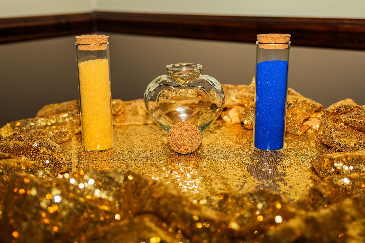 Unity sand ceremony setup with yellow and blue sand tubes beside heart-shaped glass vessel on gold glitter display.