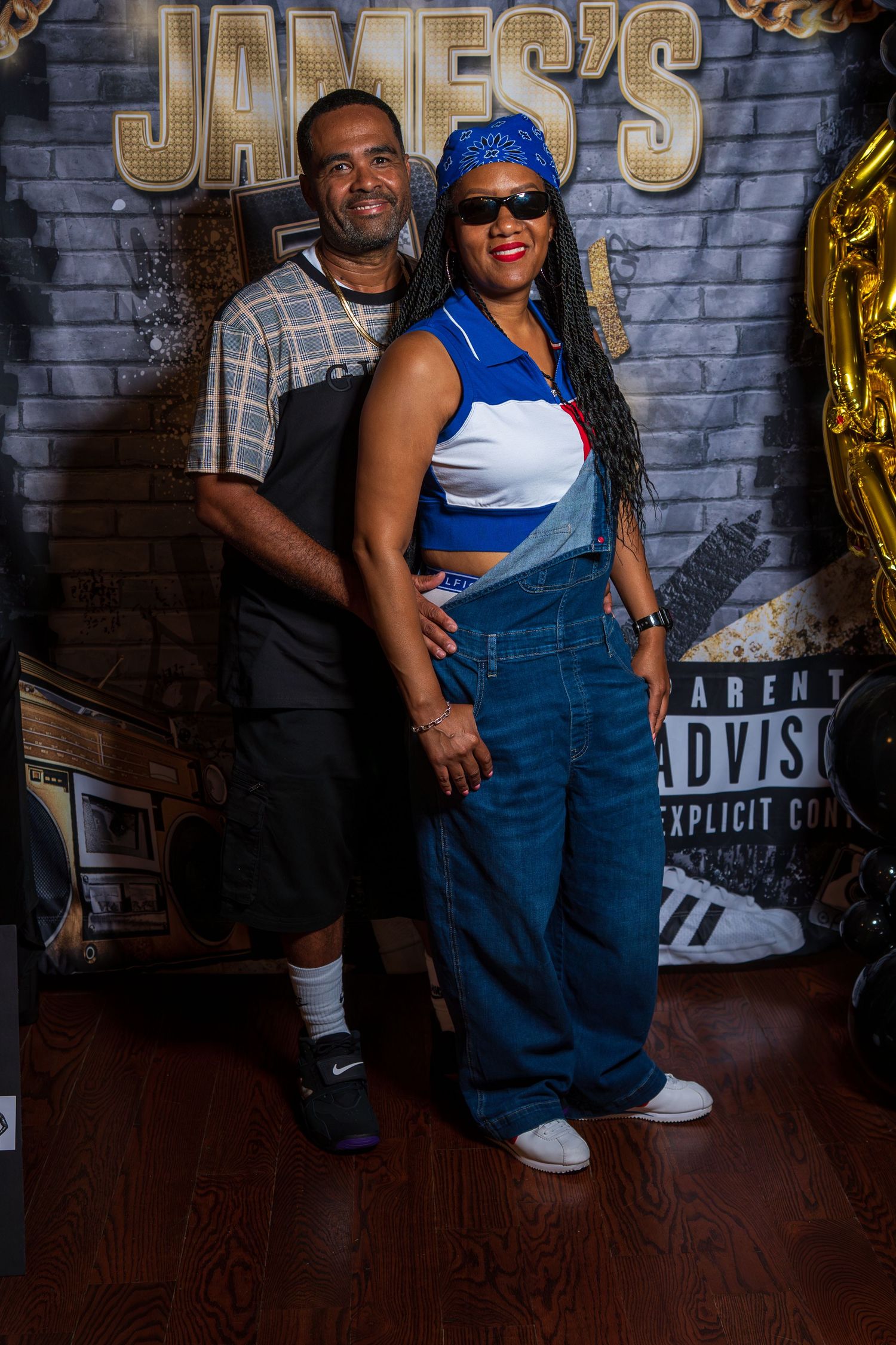 Couple posing in front of James's 50th birthday backdrop, woman in overalls and blue bandana, man in plaid shirt.