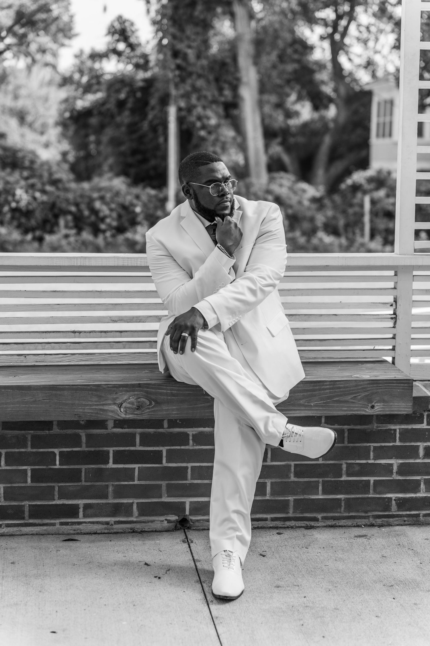 Elegant man in white suit sitting thoughtfully on bench in park setting.