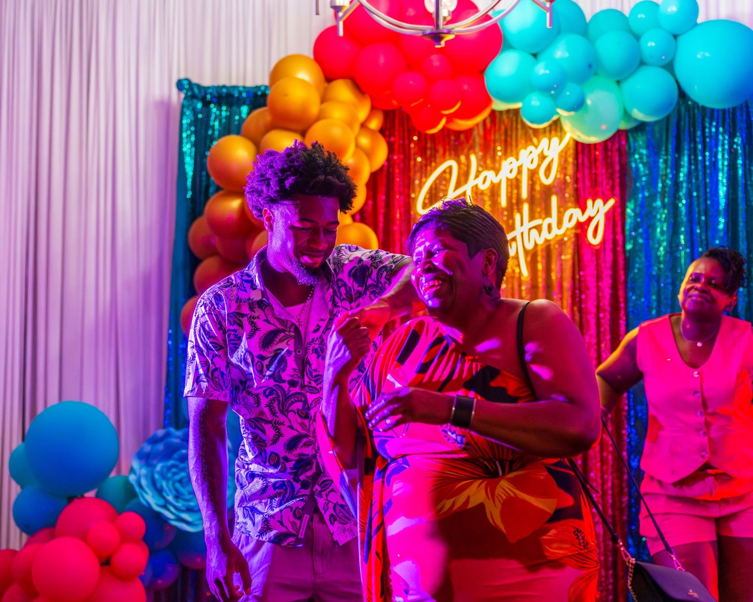 Two people dancing together under colorful balloons and a Happy Holiday neon sign at a party.