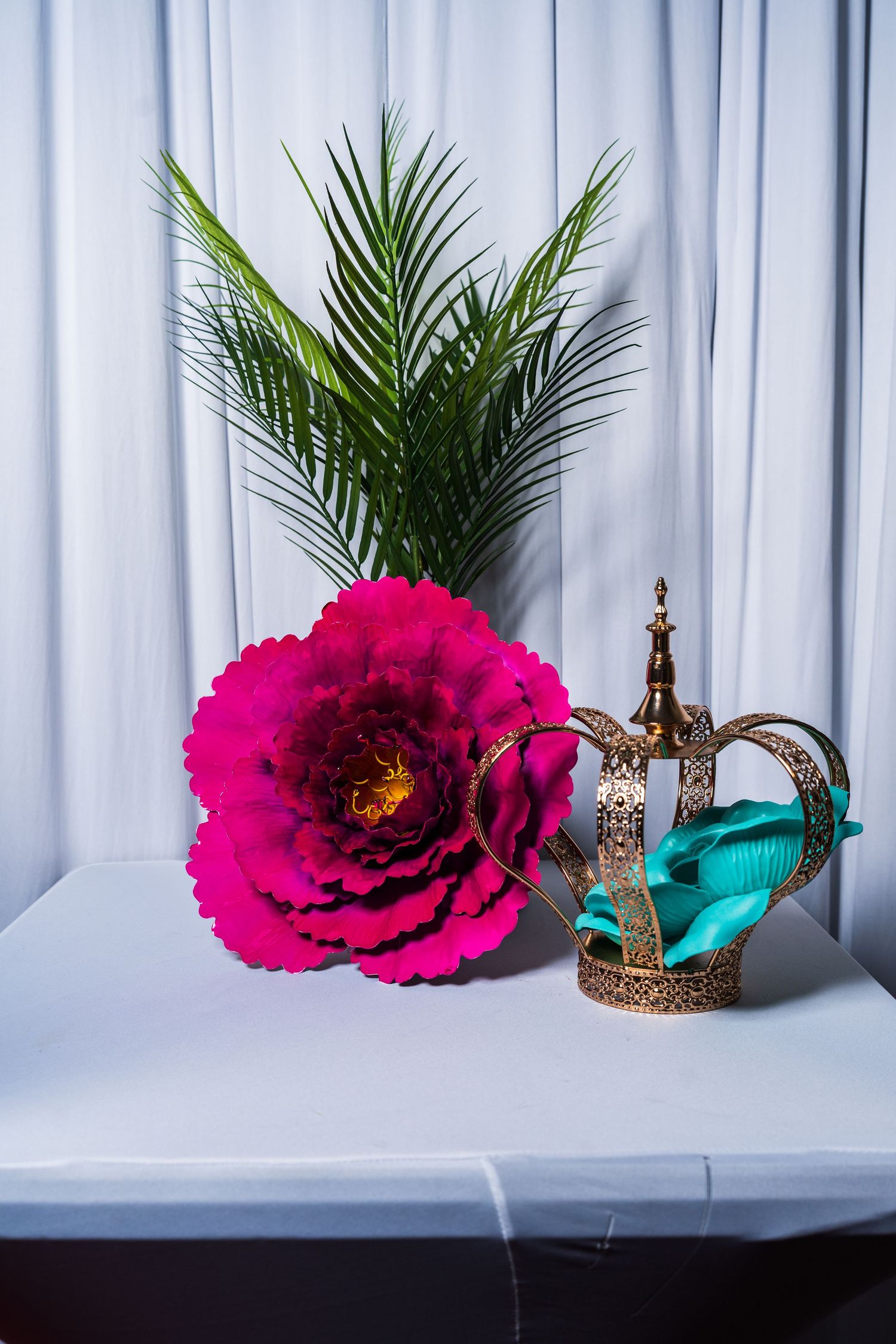 Decorative display with a large pink paper flower, gold crown, and tropical palm leaves.