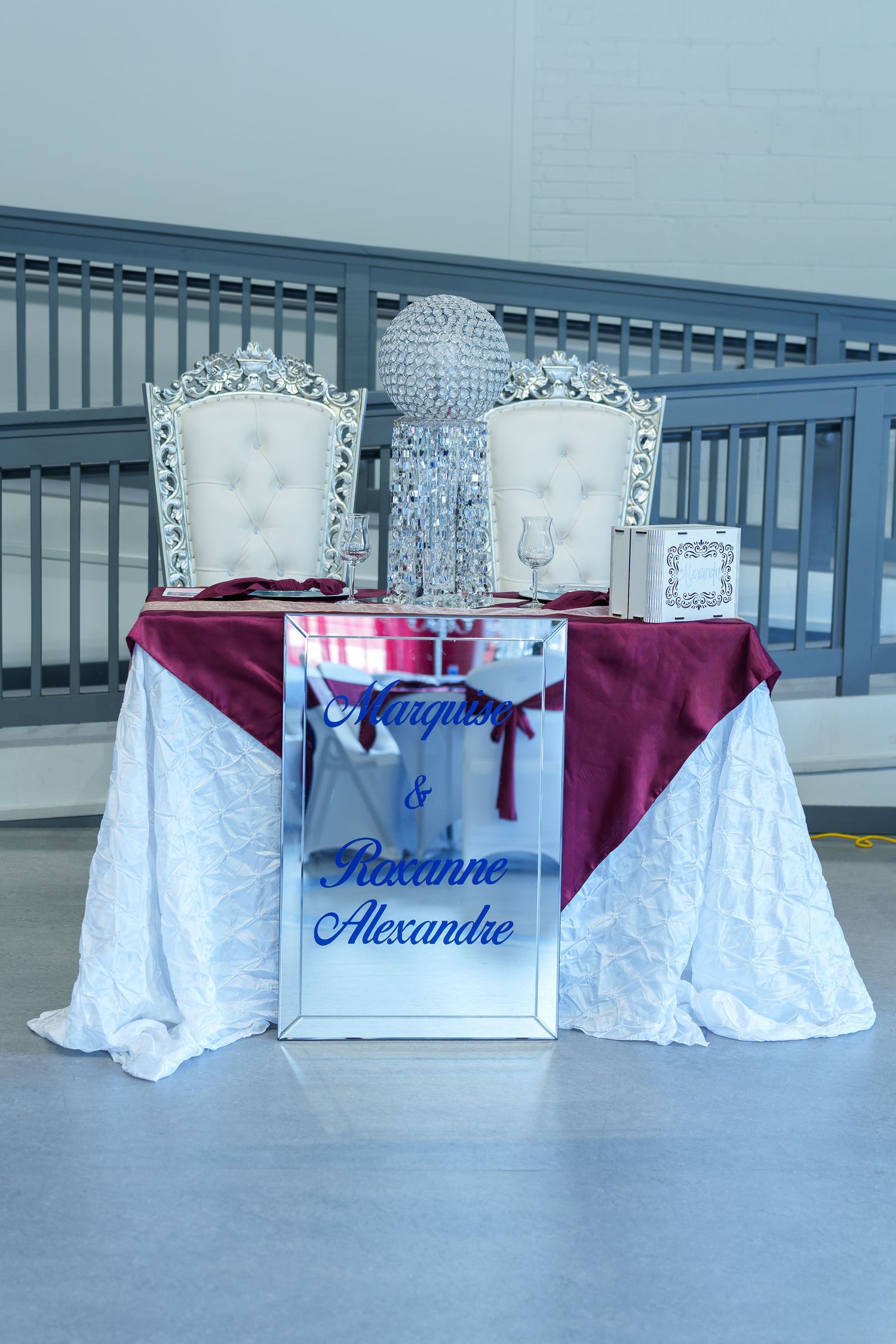 Elegant wedding sweetheart table with white tufted chairs, burgundy linens, and personalized mirror sign.