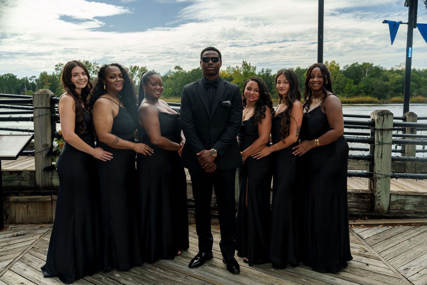 Groom in black suit poses with six bridesmaids in black gowns on a scenic waterfront dock.