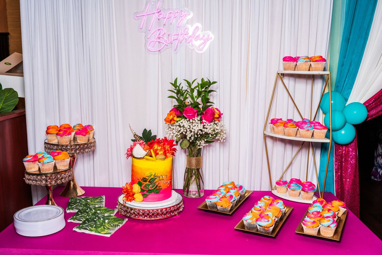 Colorful tropical birthday dessert table with cake, cupcakes, and Happy Birthday neon sign.