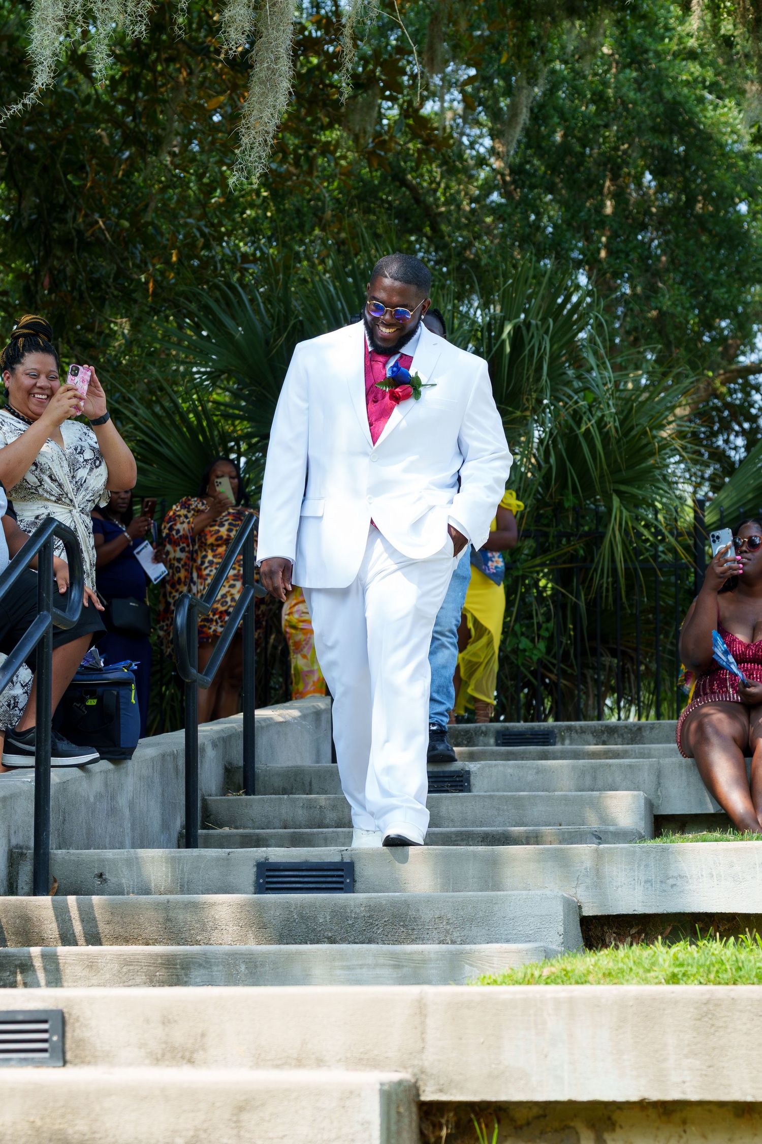 Handsome groom in white suit with pink bow tie walking up stone steps at outdoor ceremony.