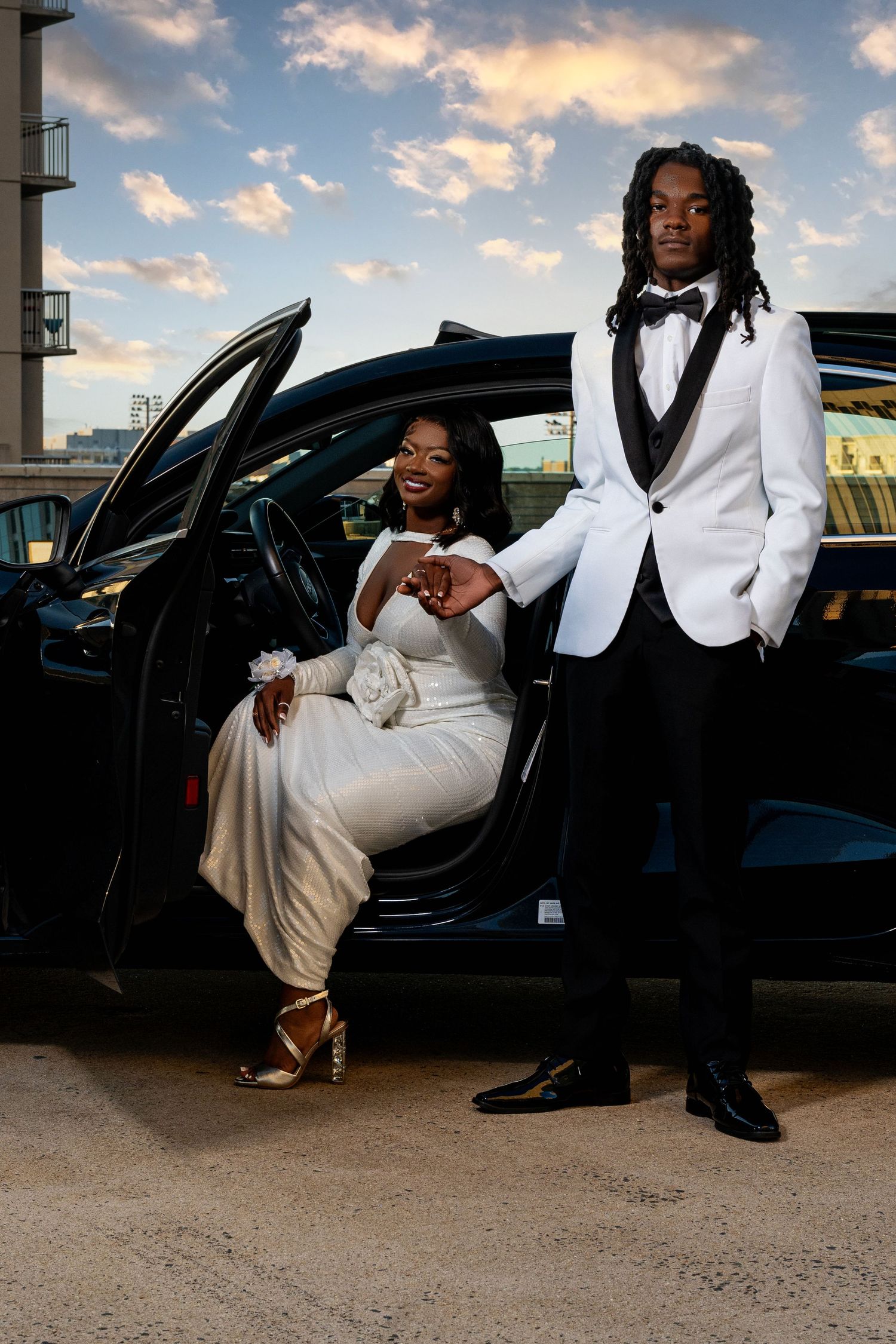 Smiling prom couple in white formal wear posing with black luxury car at sunset.