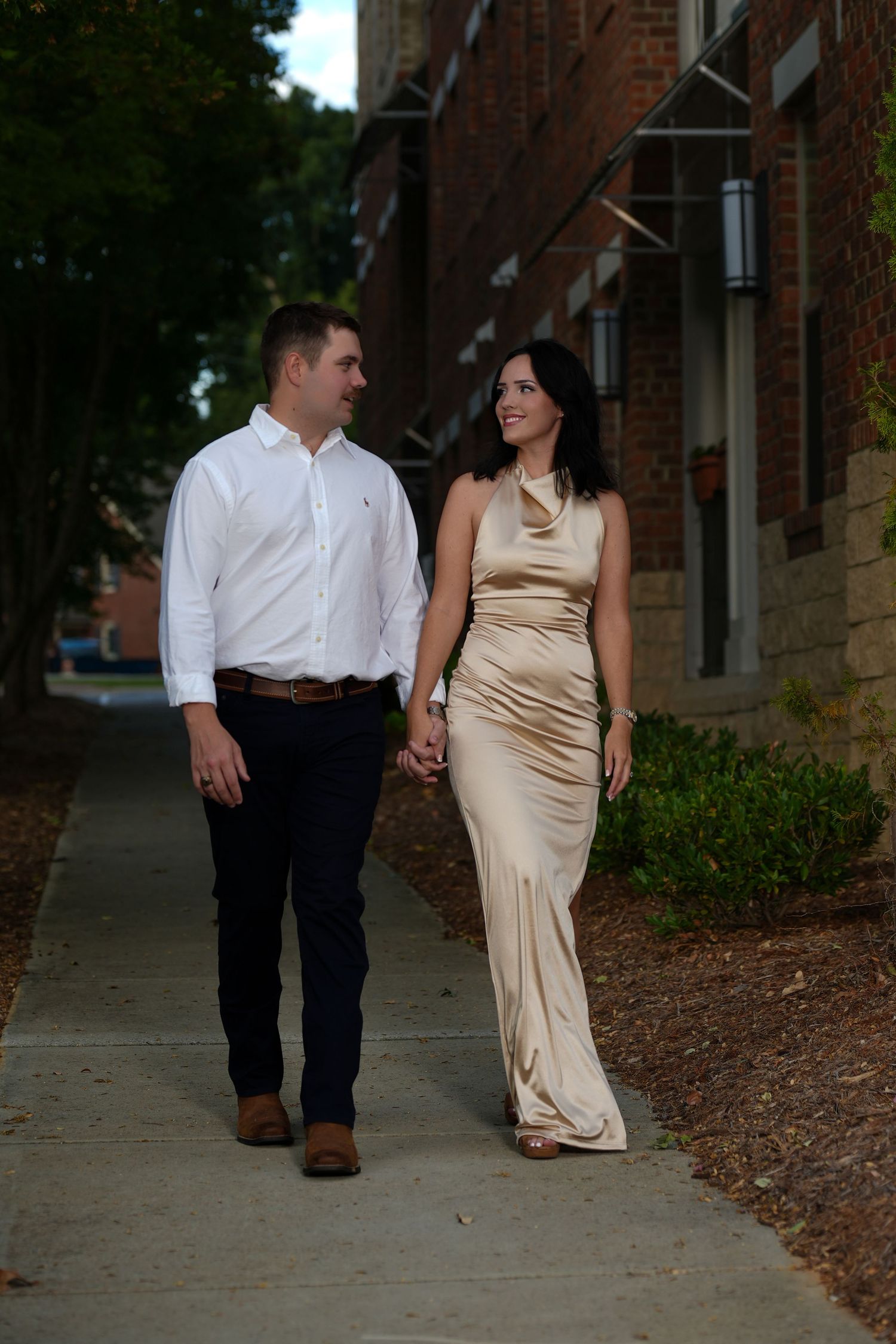 Engaged couple walking hand in hand along a brick building sidewalk.