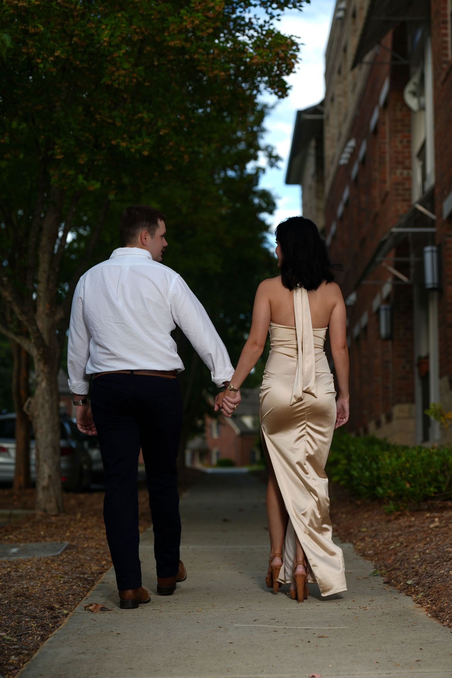 Couple walking away hand in hand on a tree-lined sidewalk near brick building.