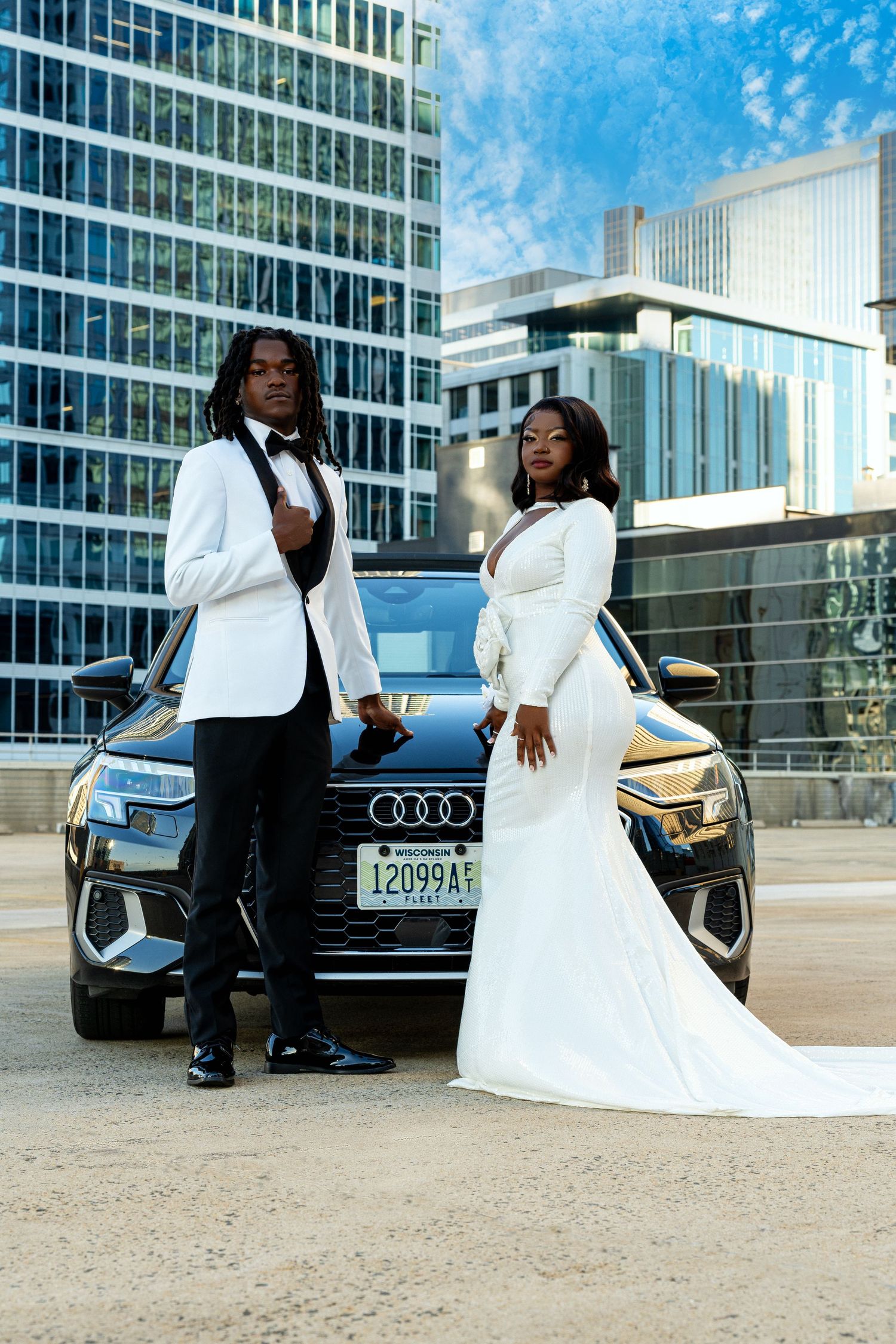 Elegant prom couple in white and black formal wear posing before Audi SUV with city backdrop.