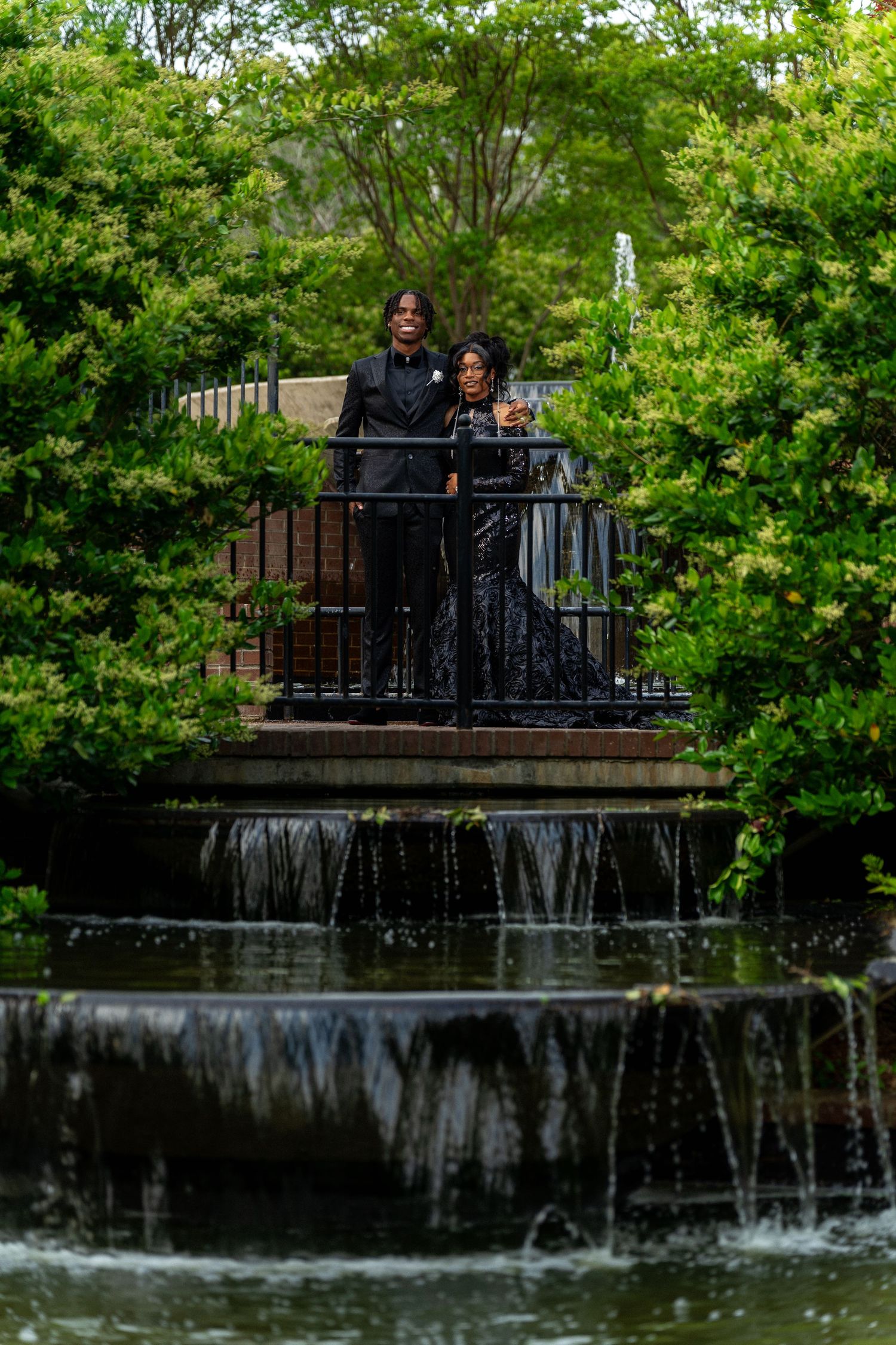 Prom couple in matching black formal wear posing on bridge over waterfall in lush garden.
