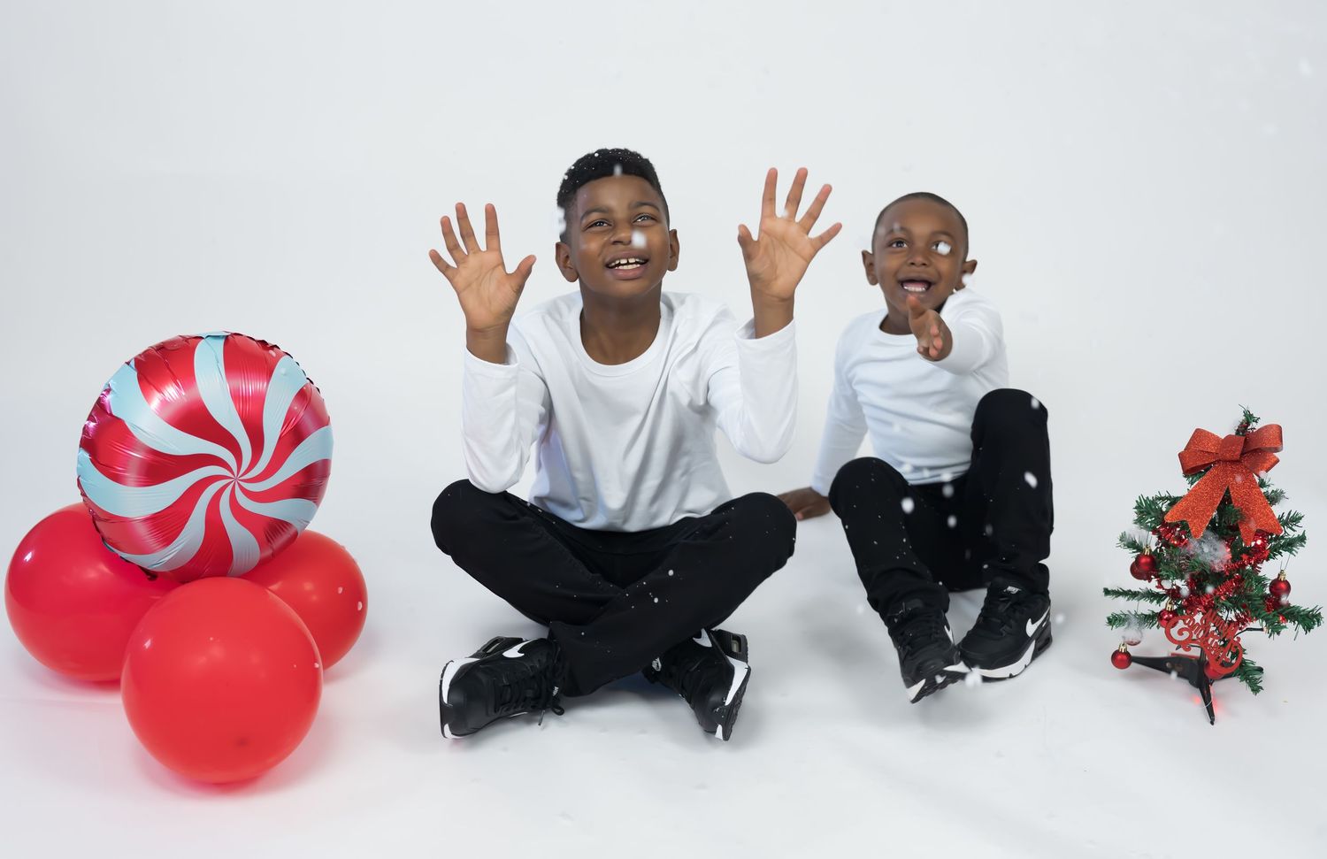 Two boys in white shirts laugh and wave hands beside red balloons and a small Christmas tree.