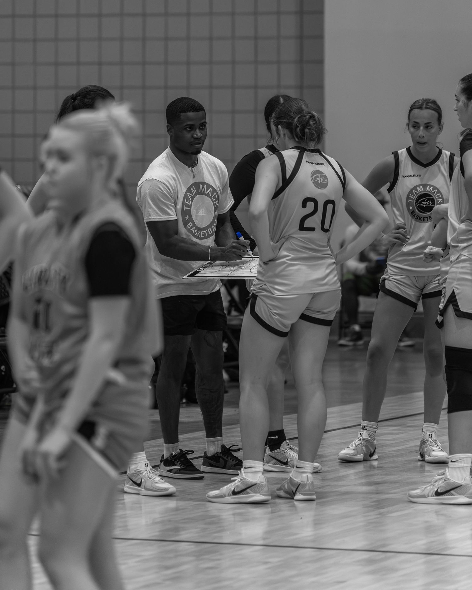 Black and white photo of a basketball coach drawing up plays on a clipboard during a timeout with female players.