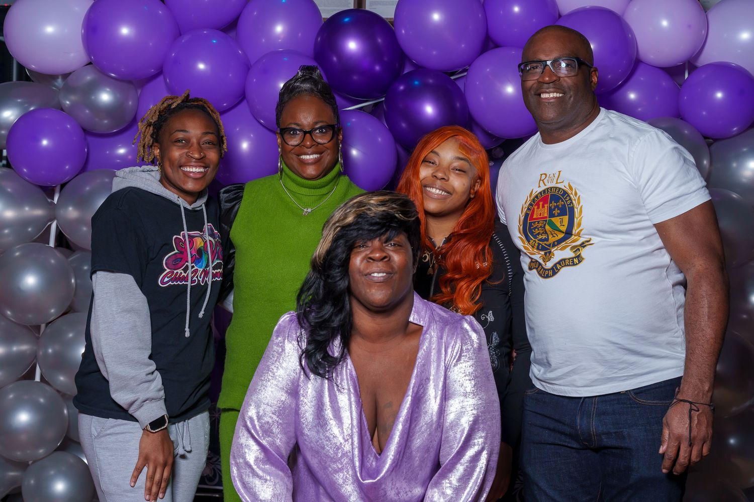 Group of five people posing in front of purple and silver balloon decorations at a celebration.