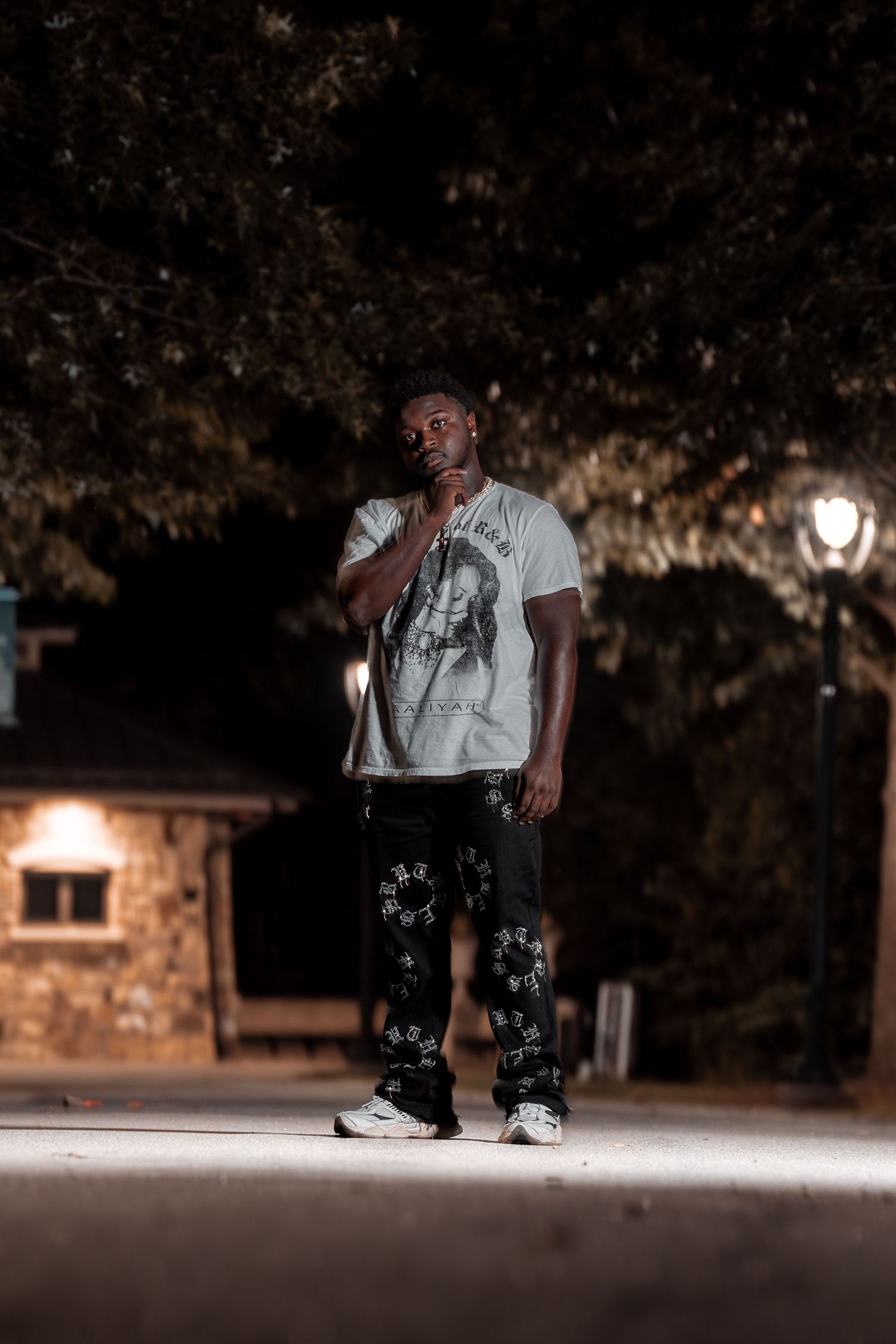 Man in white graphic tee poses thoughtfully on illuminated pathway at night near brick building and trees.