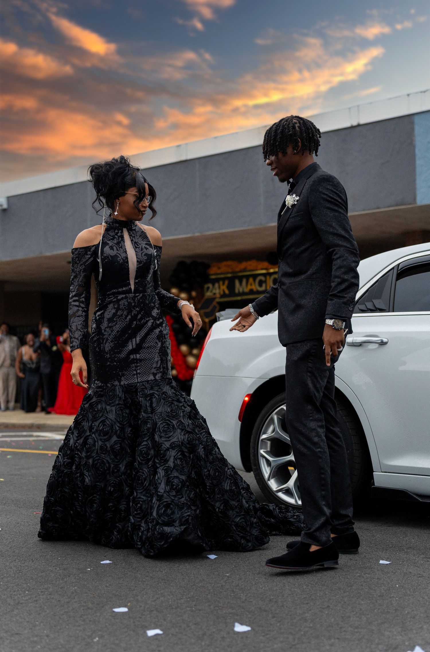 Couple in matching black formal wear arriving at prom venue at sunset from white luxury car.