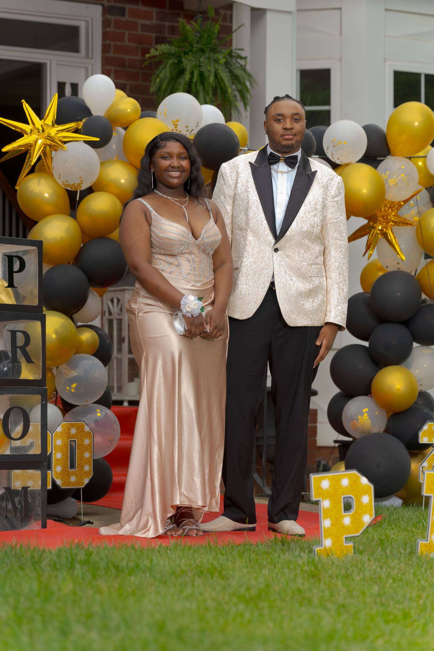 Prom couple standing before gold black and white balloon arch with PROM marquee letters on red carpet.