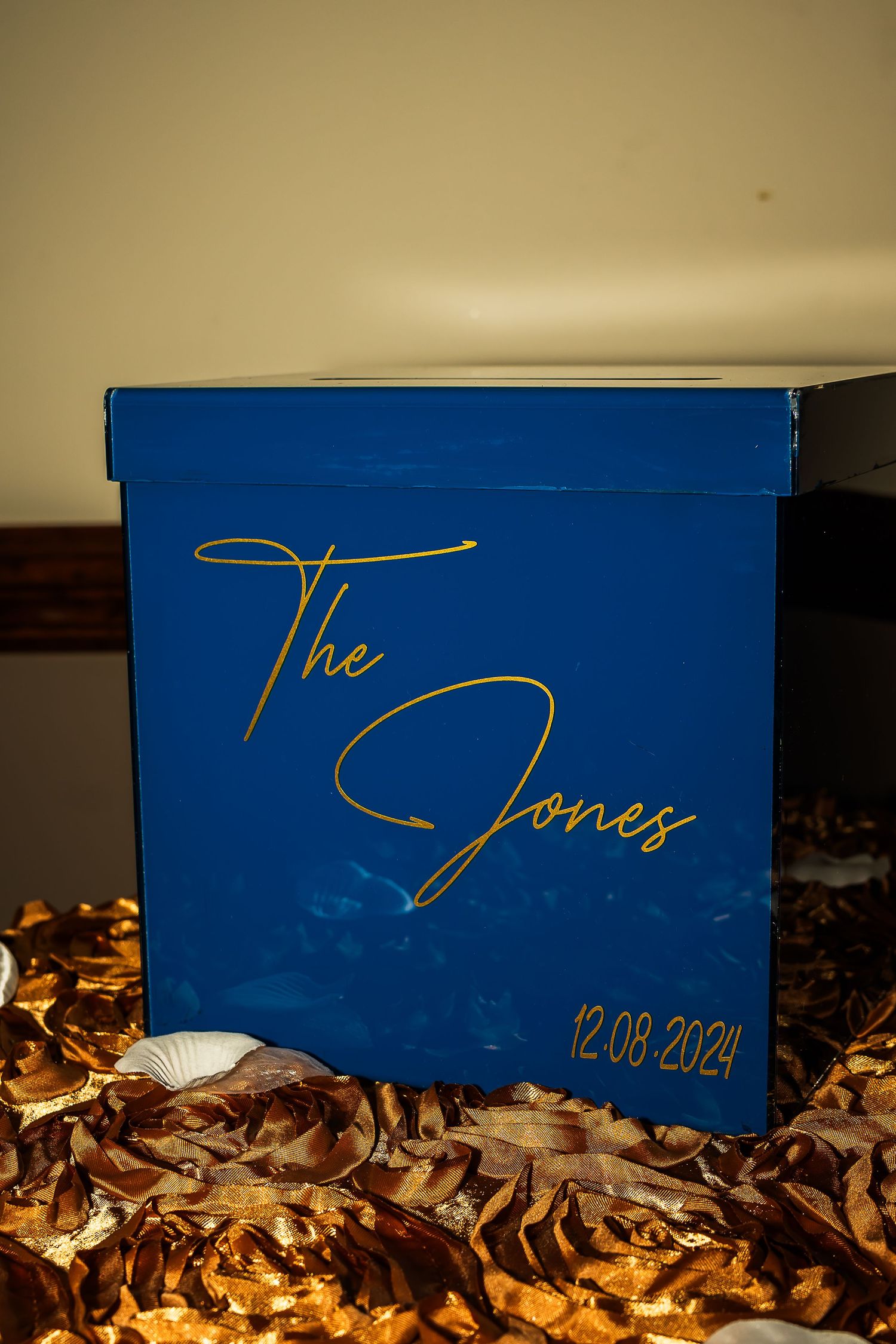 Royal blue wedding card box with gold script reading The Jones dated 12-08-2024, surrounded by gold decorations.