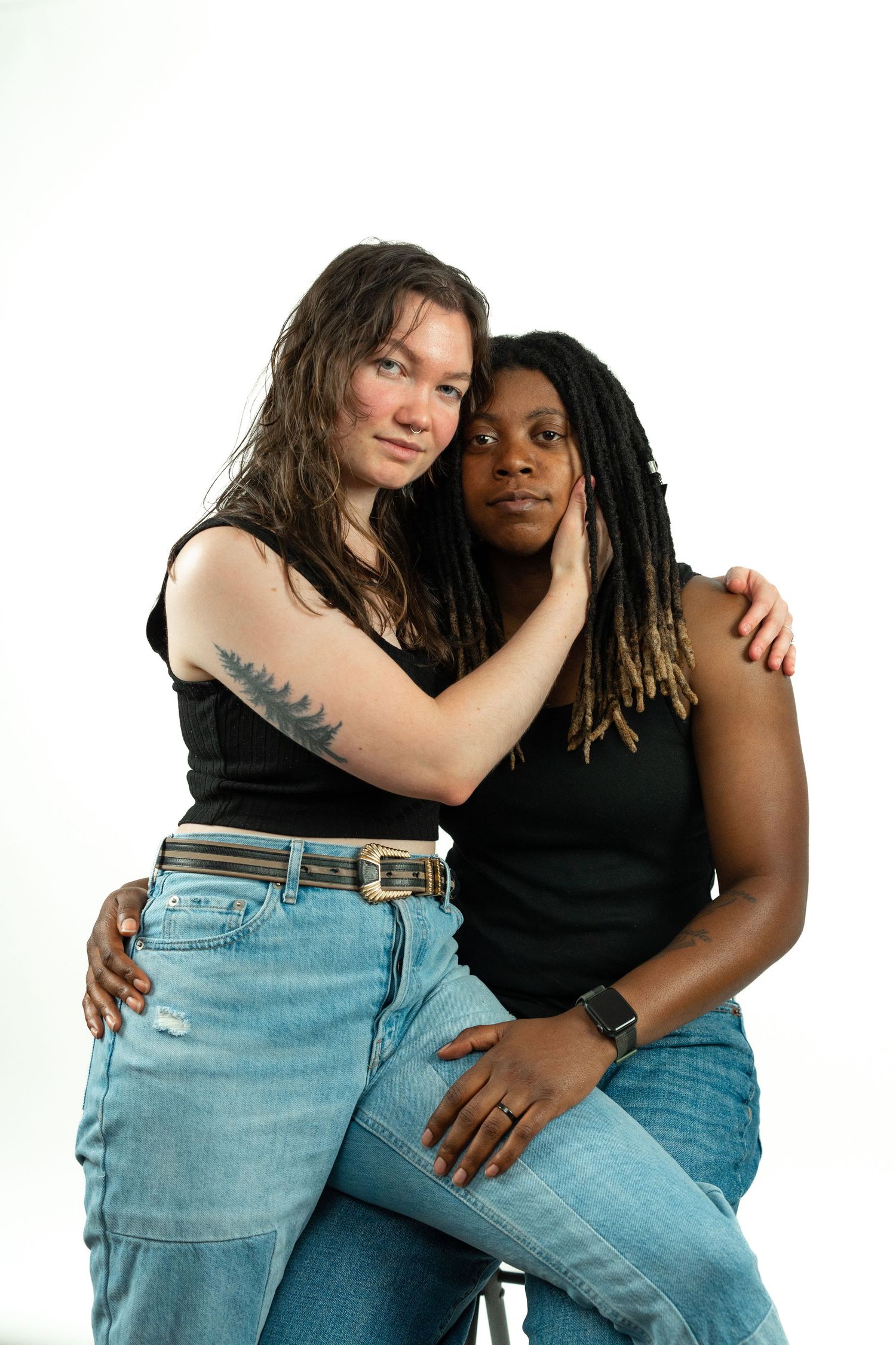 Two women embracing in a studio portrait, one with tattoos wearing jeans and black top.