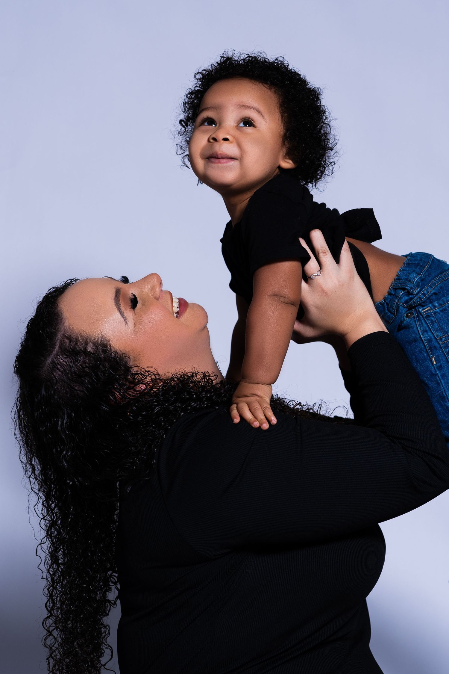 Joyful mother lifts laughing toddler in black outfit against light gray studio background.