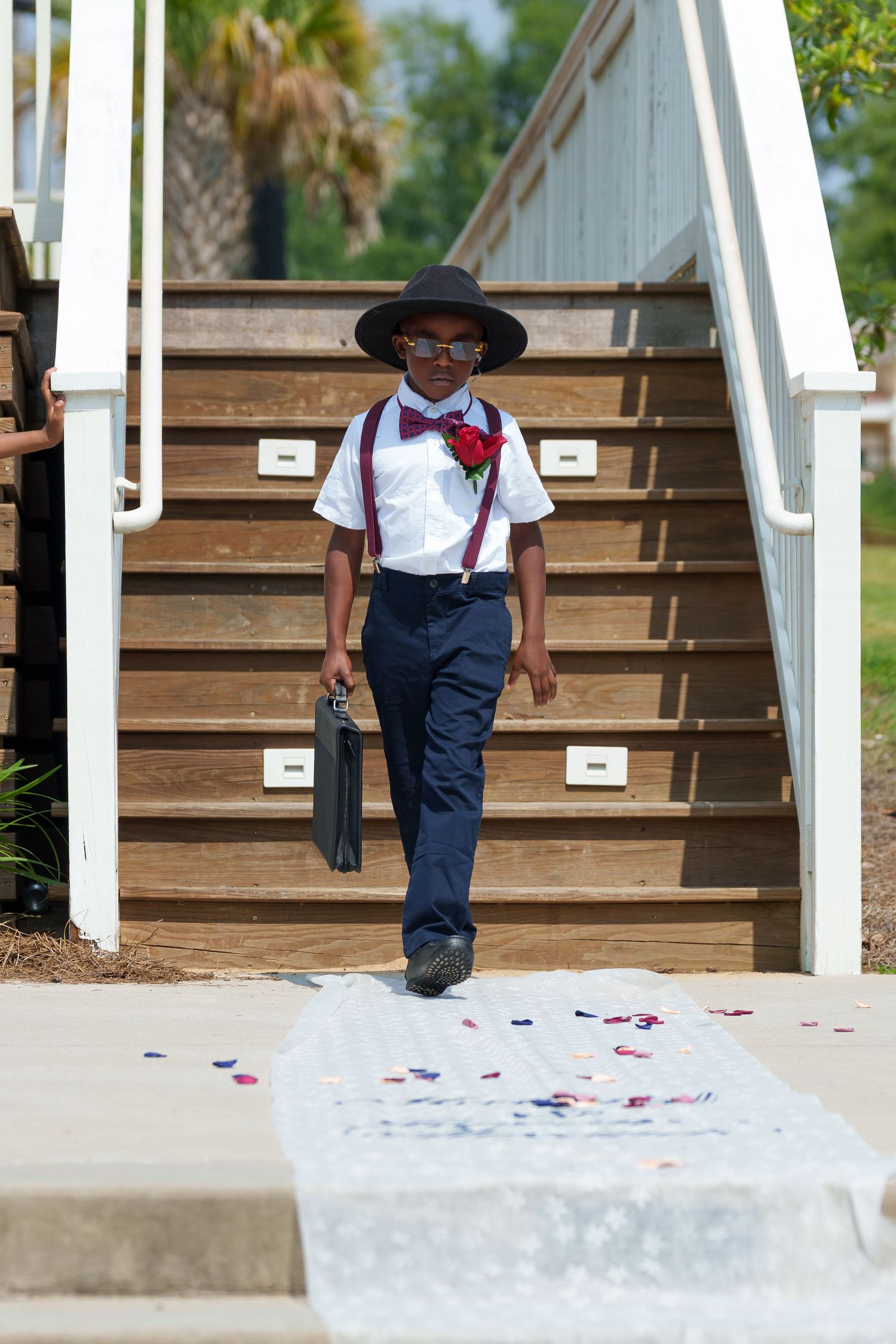 Stylish young ring bearer in hat, bow tie, and suspenders walking down wedding aisle.