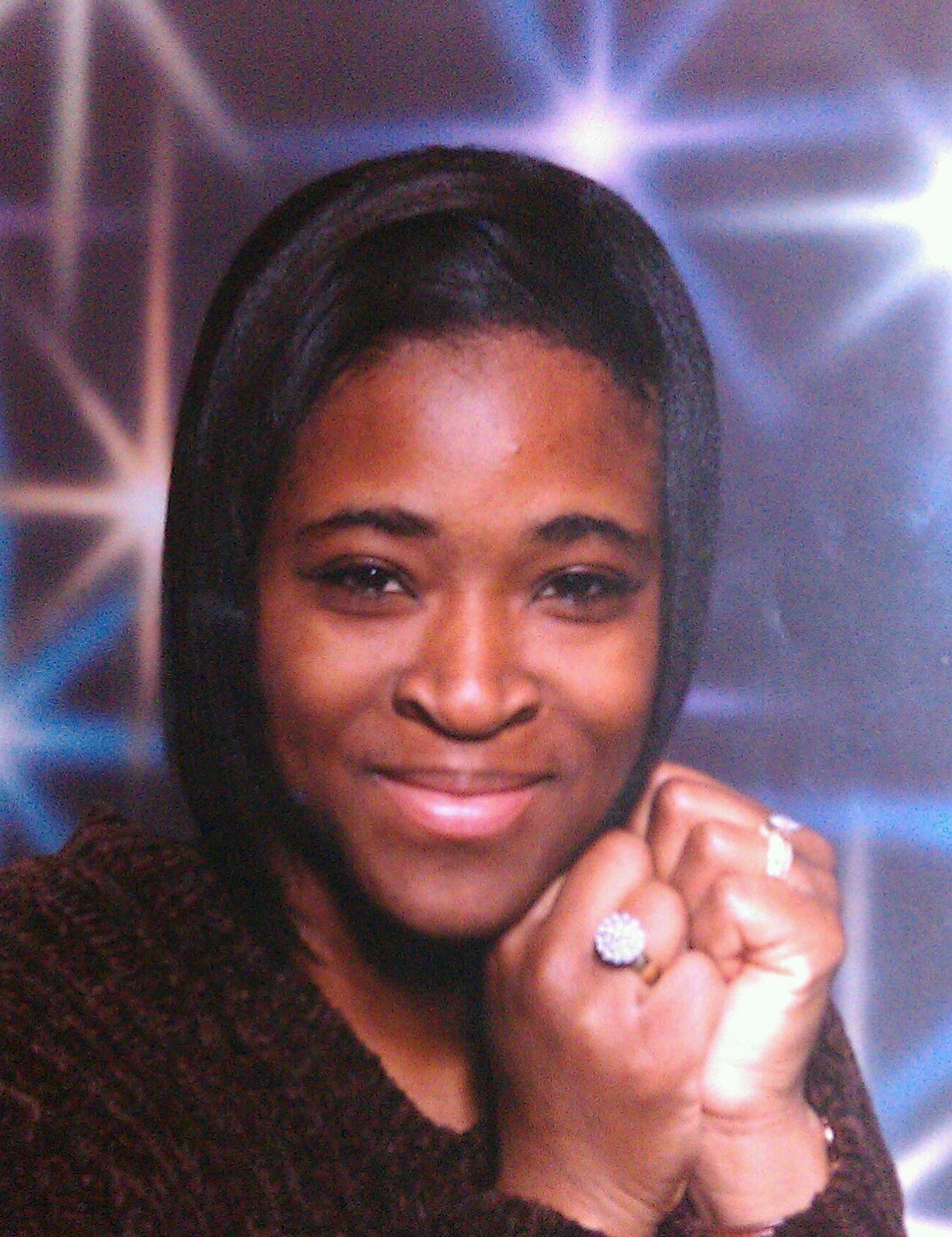 A smiling woman poses with hands clasped under her chin against a blue starry background in a portrait.