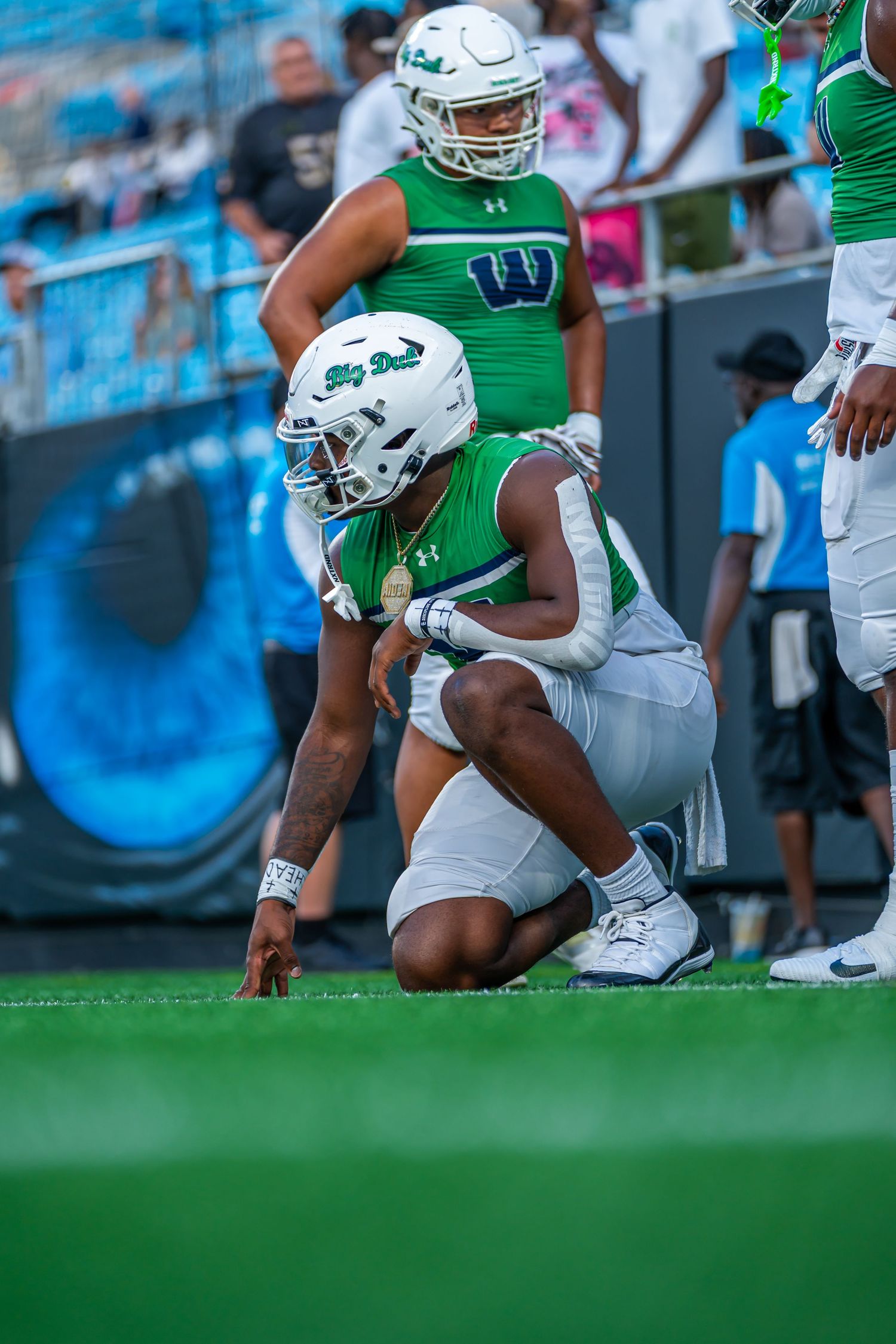 Football player in green and white uniform crouches in ready position on game field.