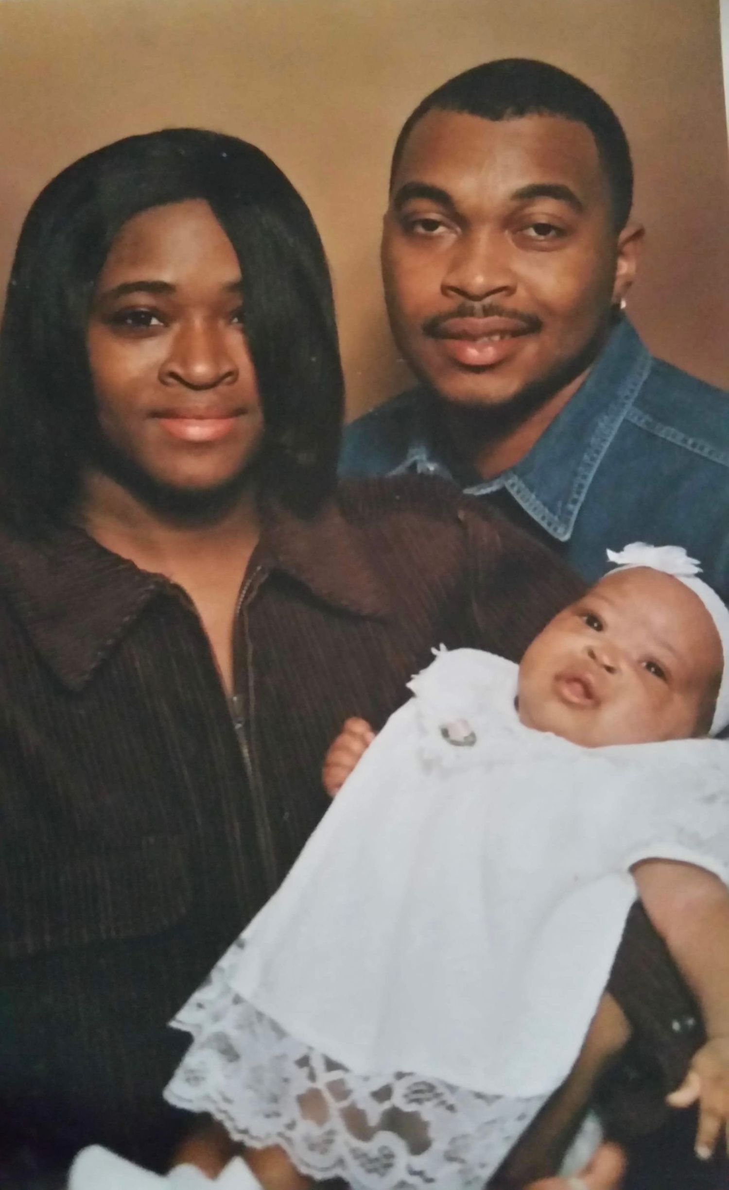 Family portrait of a couple in brown and denim outfits holding their infant dressed in white lace.