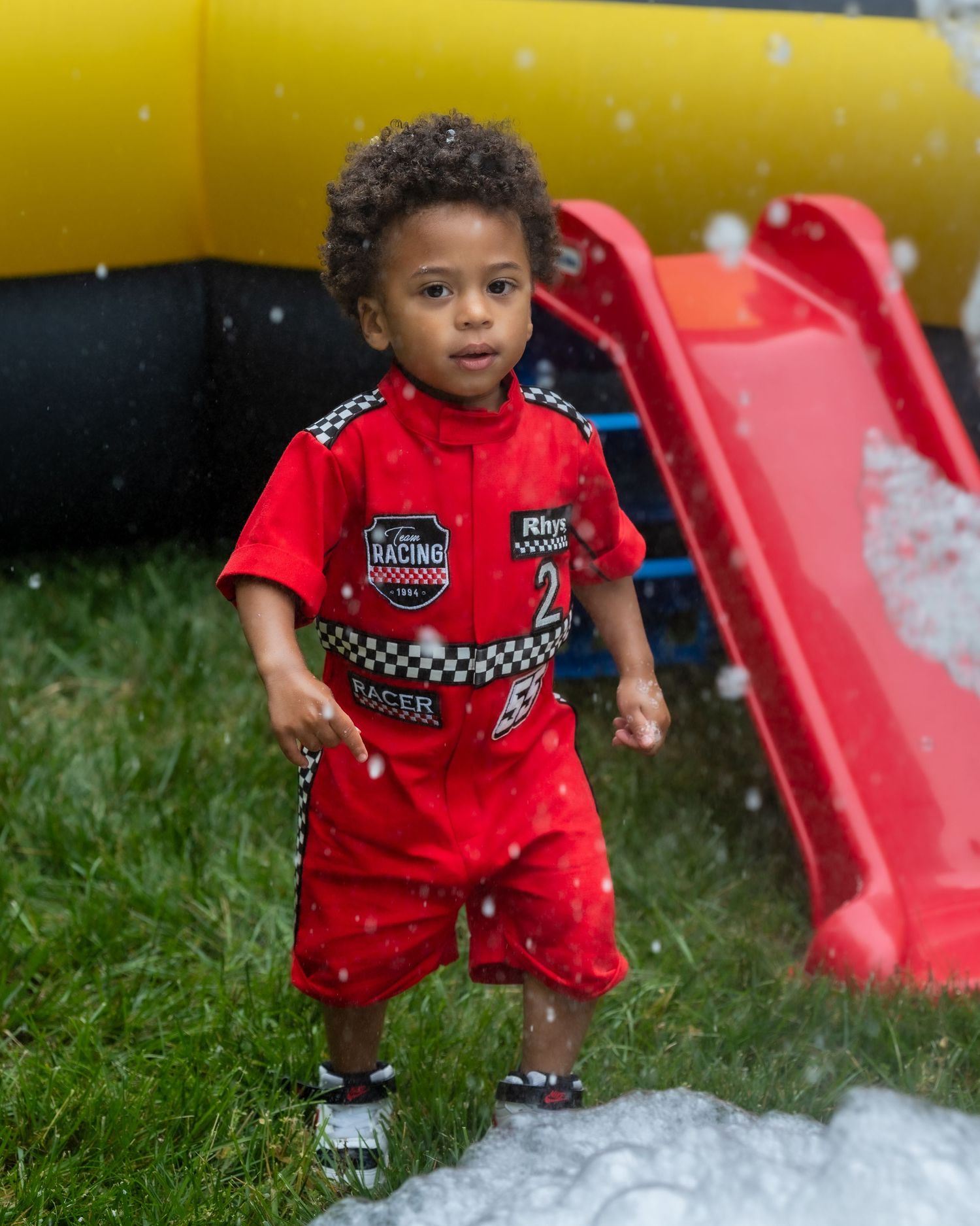 Toddler boy in red racing costume stands on grass near a red slide at outdoor birthday party.