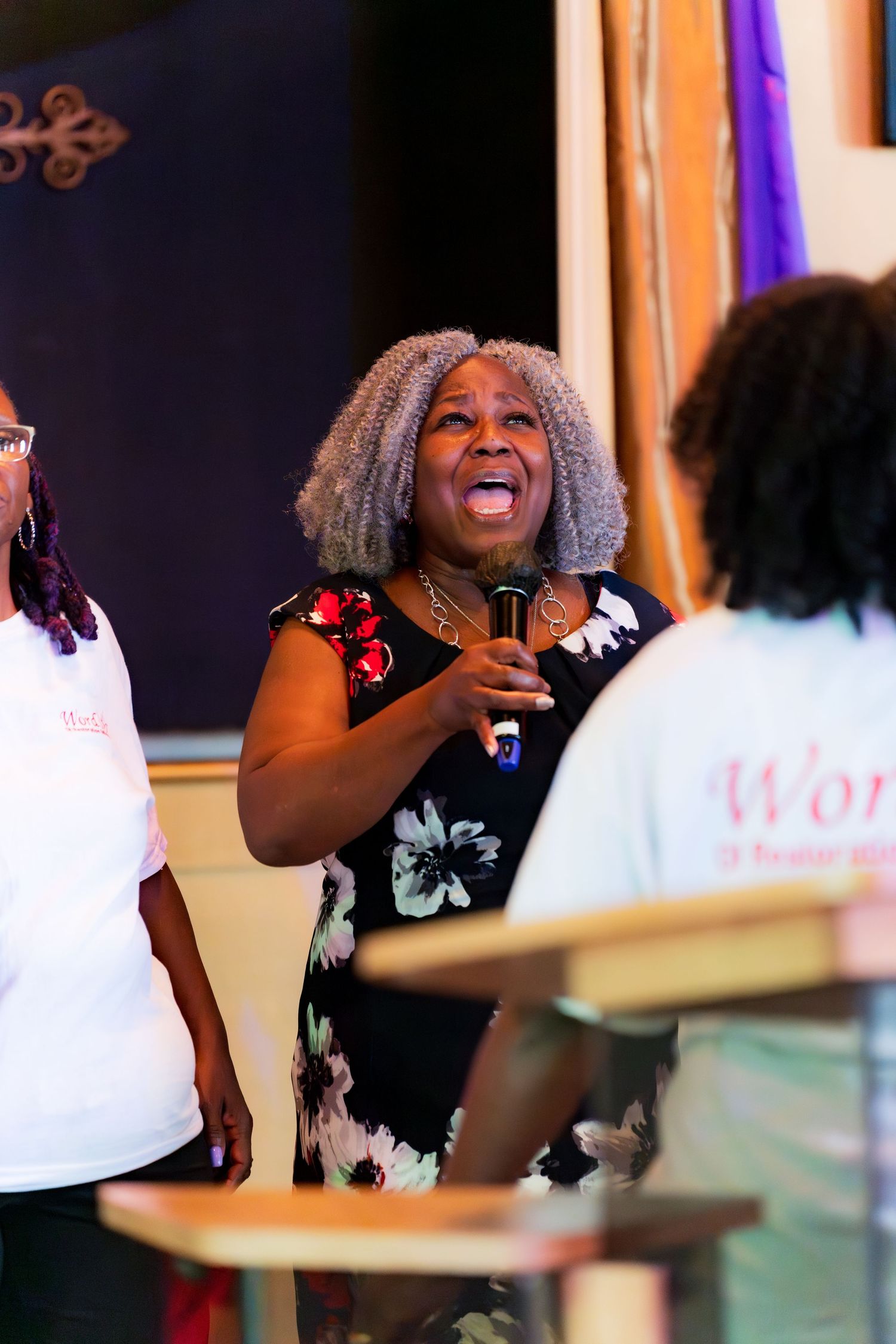 A woman with gray locs sings joyfully into a microphone at a church event.