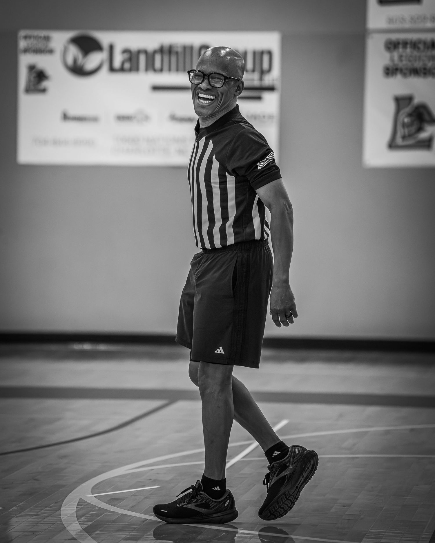 Smiling referee in black and white striped uniform walking on an indoor basketball court.