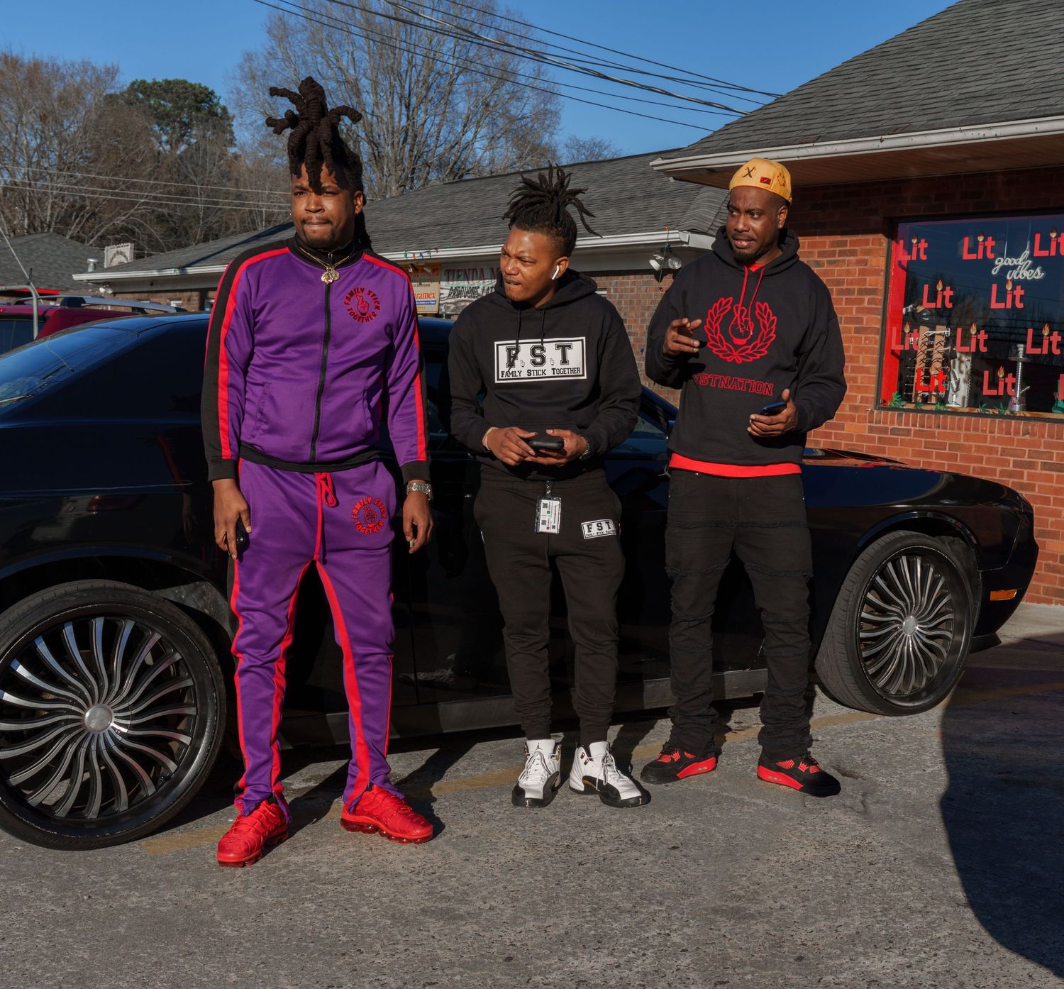 Three stylish men standing together near black car outside brick building.