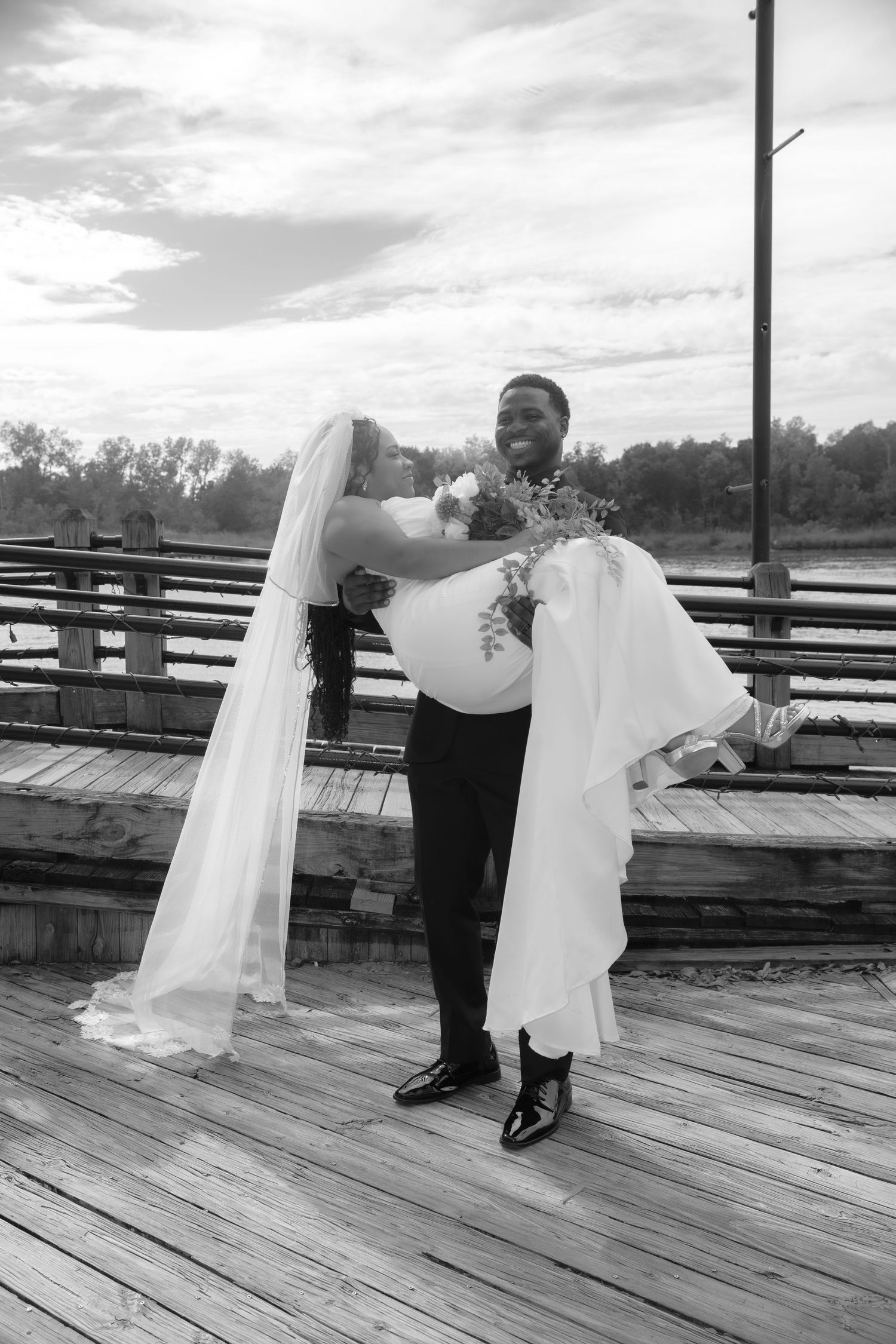 Black and white photo of groom lifting smiling bride in wedding dress on a wooden pier outdoors.