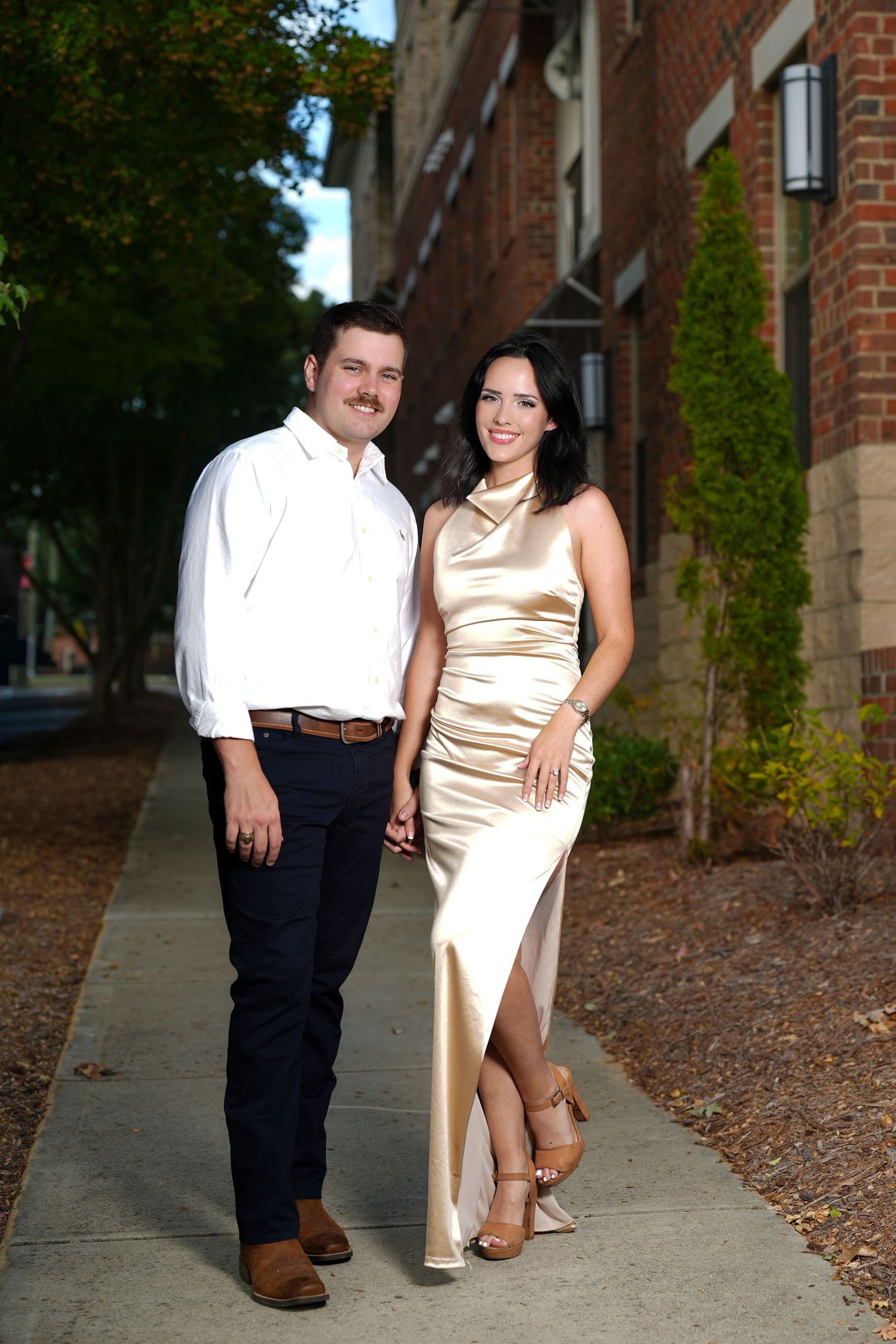 Couple posing on sidewalk, man in white shirt and woman in gold slit dress.