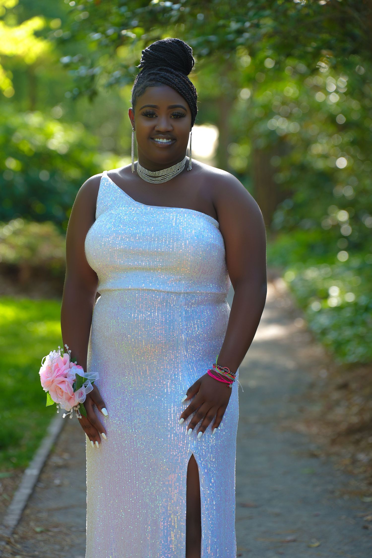 A woman poses elegantly in a white sequined one-shoulder prom gown with a pink floral wrist corsage outdoors.