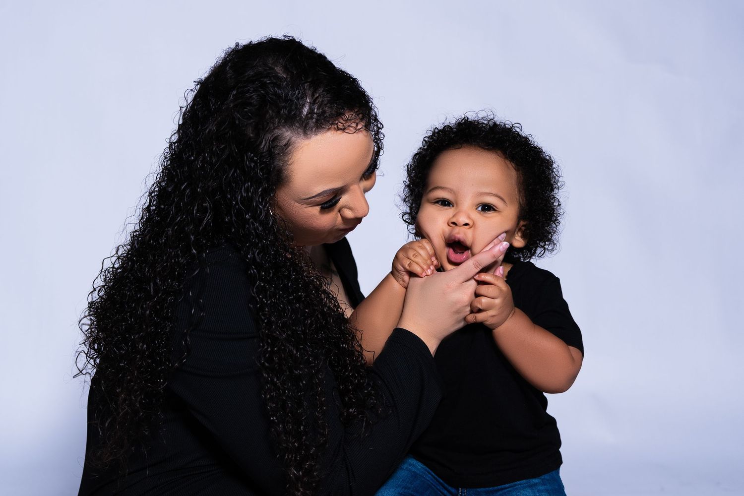 Mother with curly black hair in black outfit holds smiling baby in studio portrait.