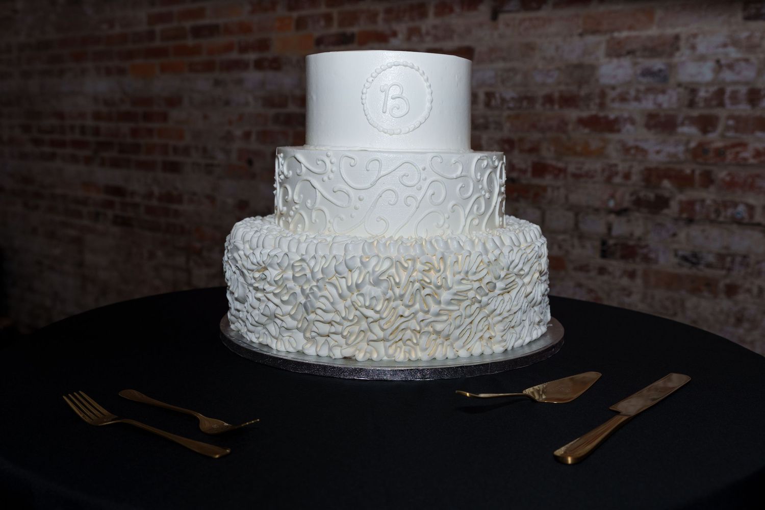 Elegant three-tier white wedding cake with intricate floral texture on a black tablecloth against brick wall.