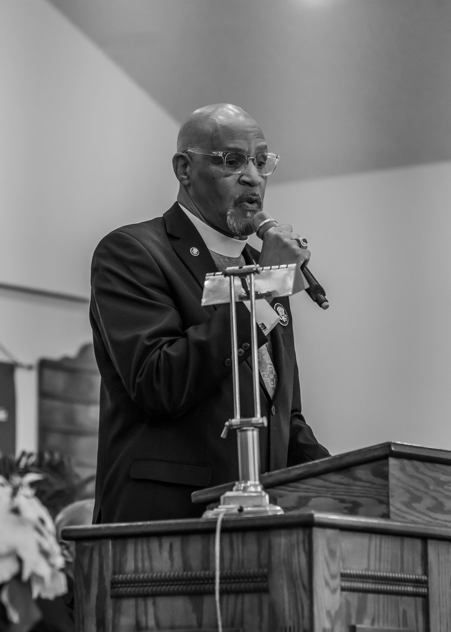 An elderly man in a black suit speaks passionately into a microphone at a church podium in black and white.