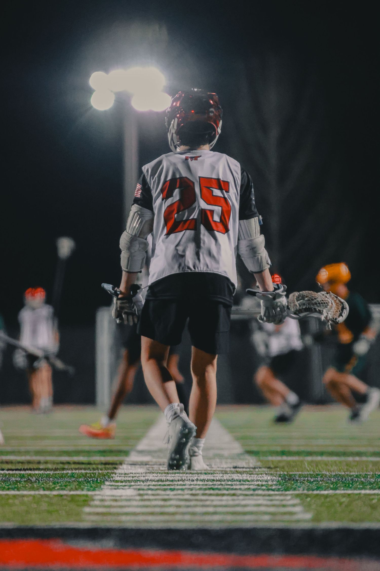 Lacrosse player number 25 walking on field under bright stadium lights during nighttime game.