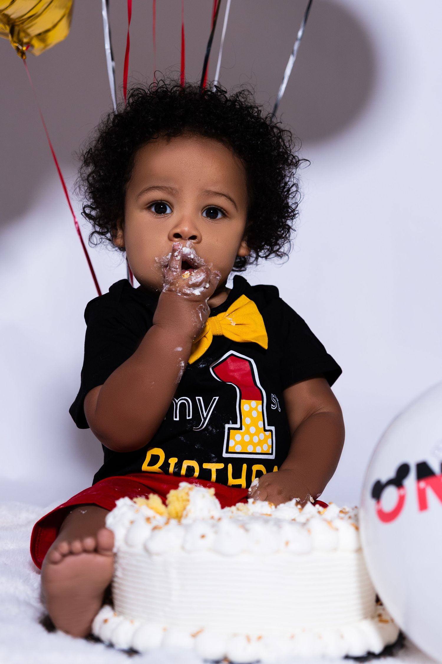 Cute baby boy eating first birthday cake wearing My 1st Birthday outfit with balloons.