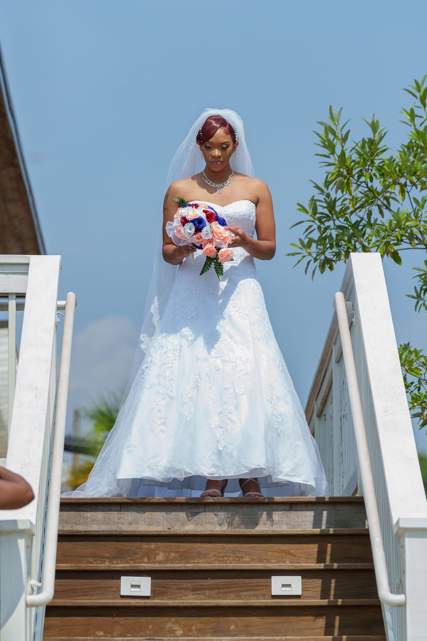 Bride in white lace gown holding bouquet standing on stairs under blue sky.