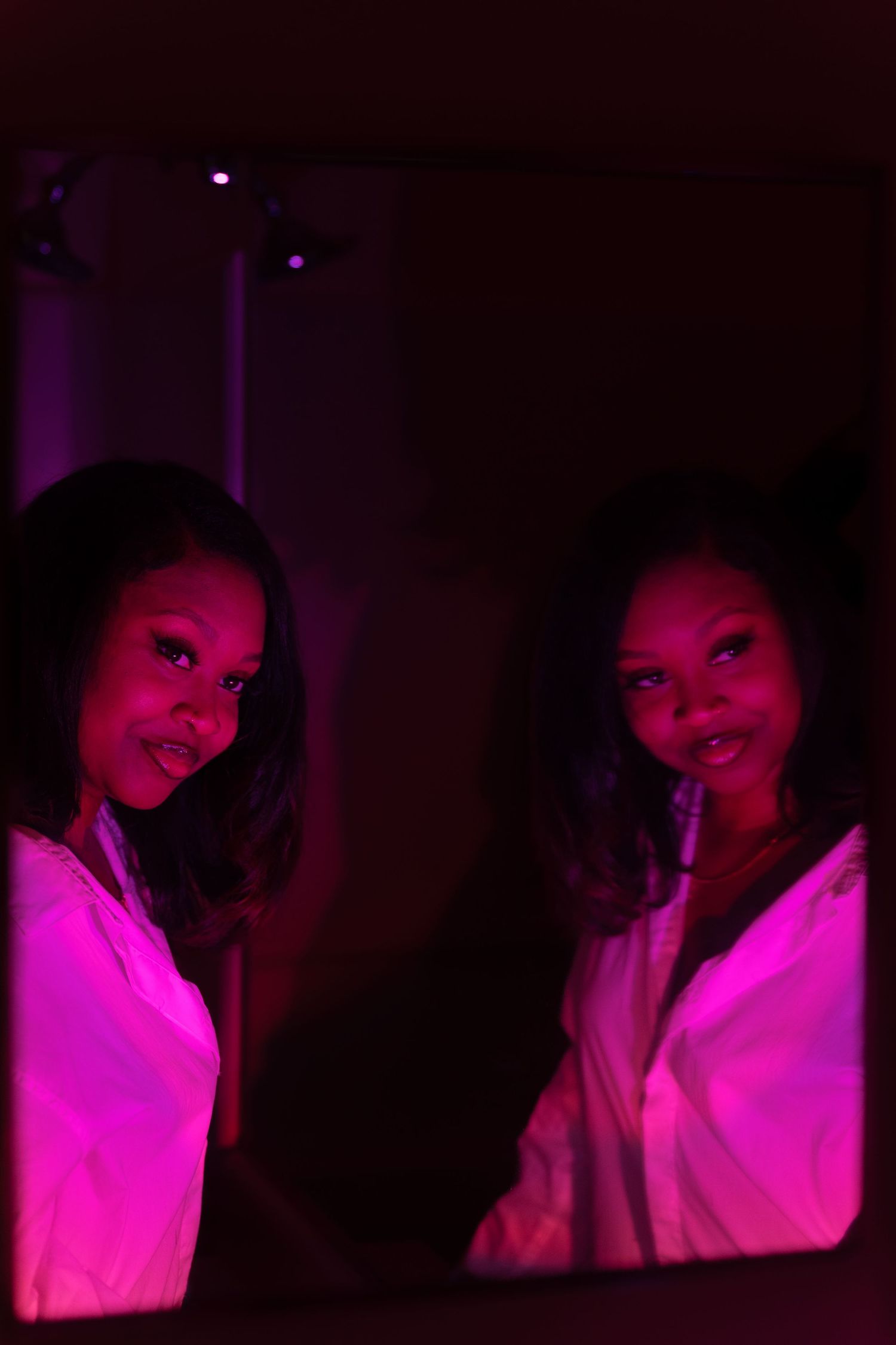 Woman in white jacket reflected in mirror under pink neon light.