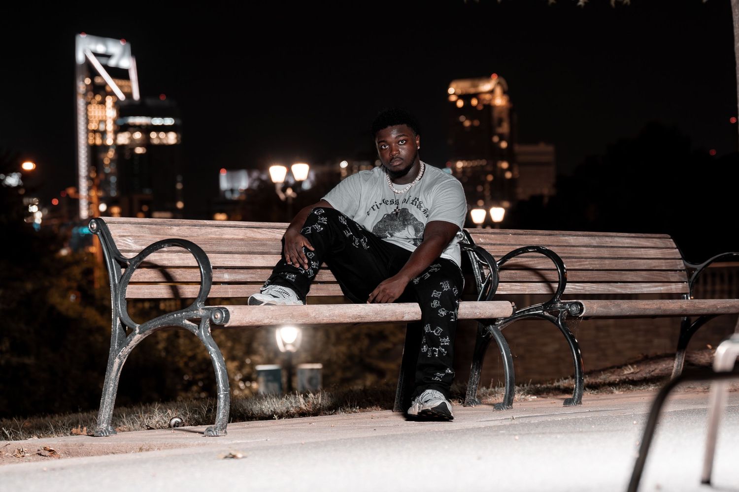Man in graphic tee and camo pants sits alone on park bench at night with city lights in background.