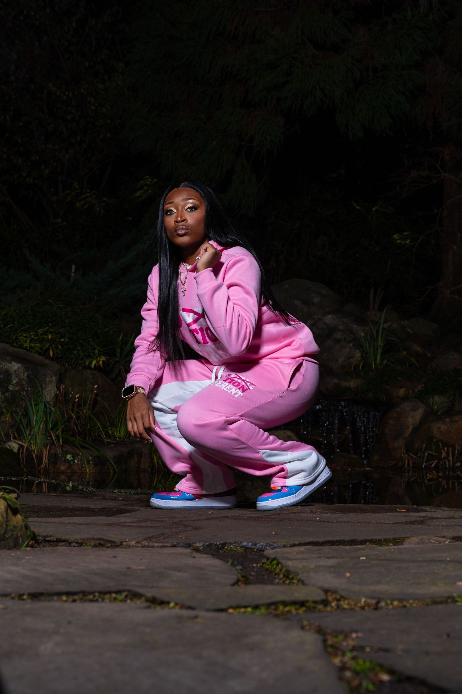 Woman in pink tracksuit and colorful sneakers crouches outdoors near rocks and greenery at night.