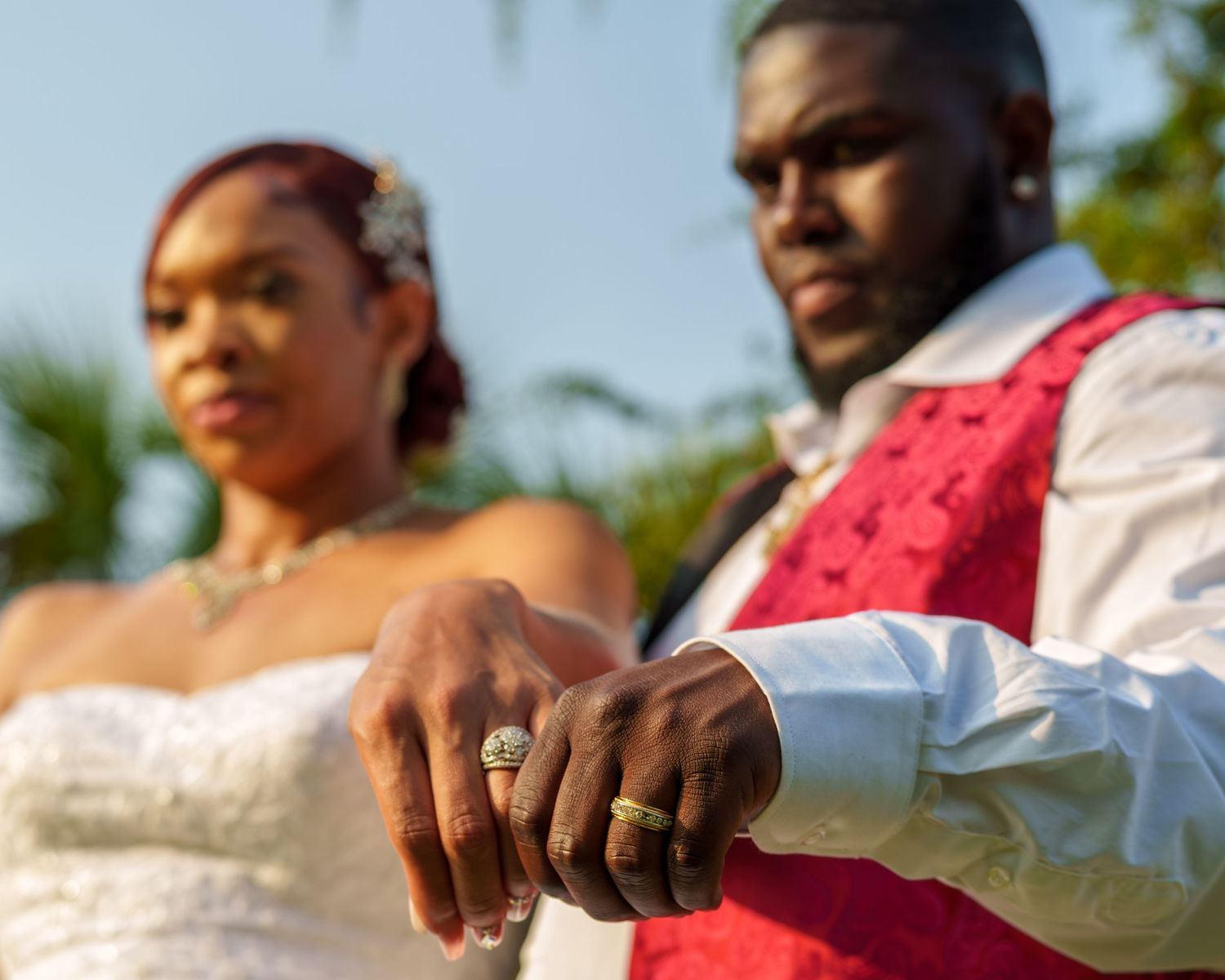 Black couple displaying wedding rings at outdoor ceremony, groom in red vest and white shirt.