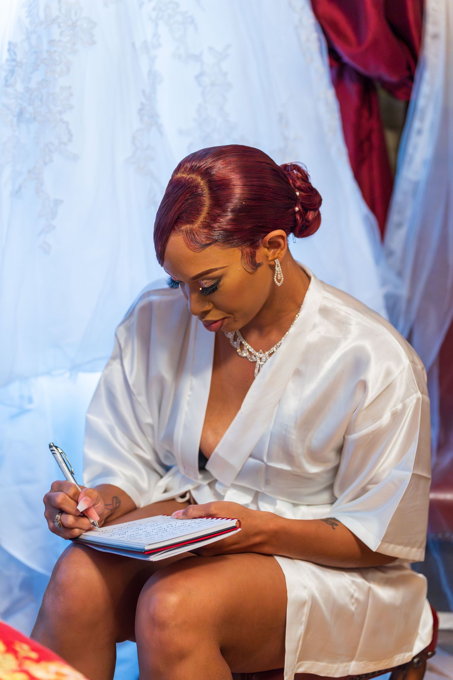 Bride in white satin robe with red updo writing vows in a notebook before her wedding ceremony.