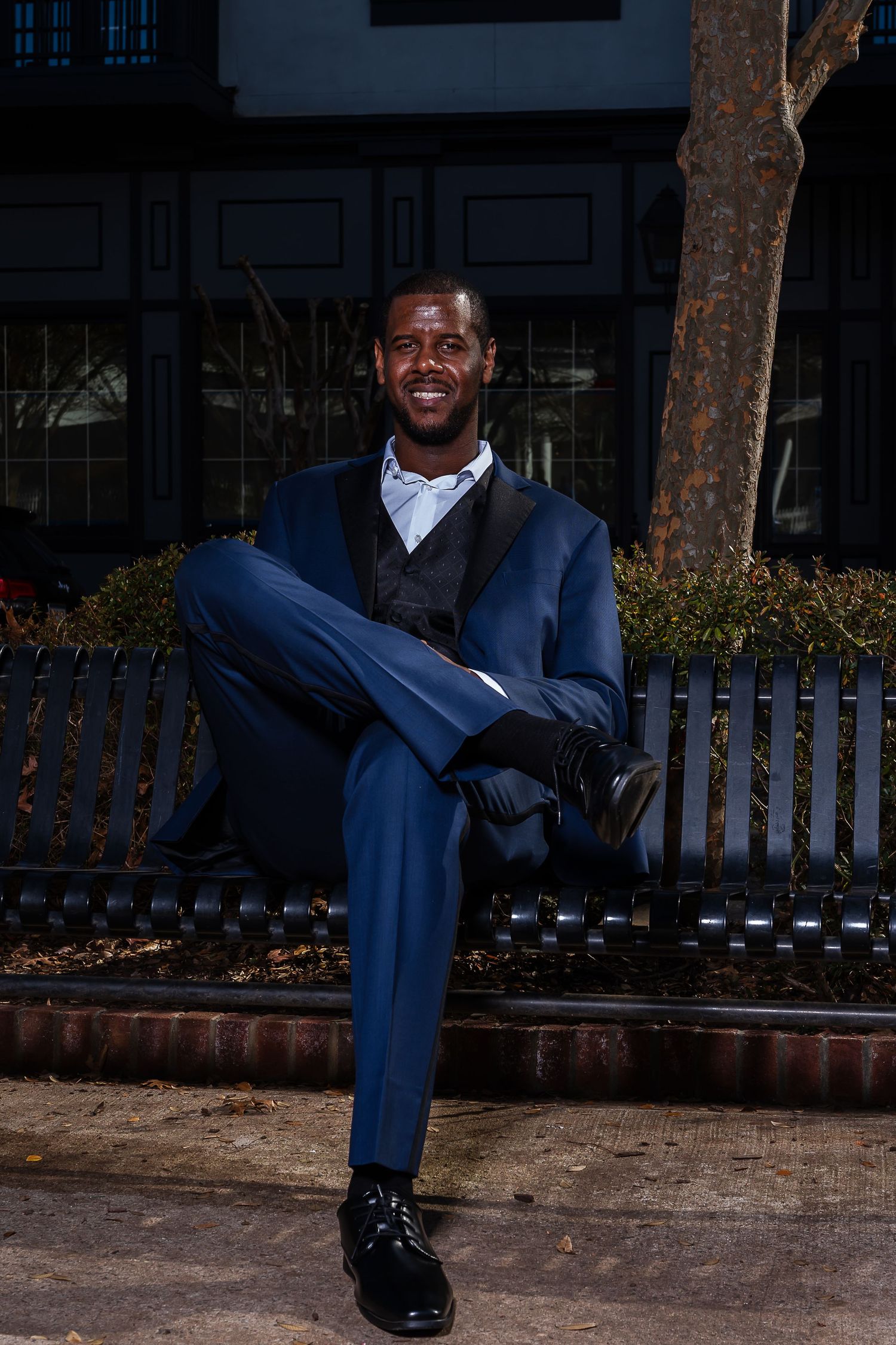 Smiling groom in navy blue suit sitting relaxed on iron bench outdoors before wedding ceremony.