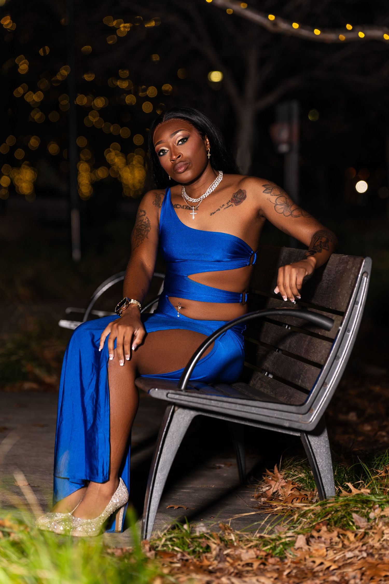 Woman in blue cutout dress sitting on bench at night with bokeh lights in background.