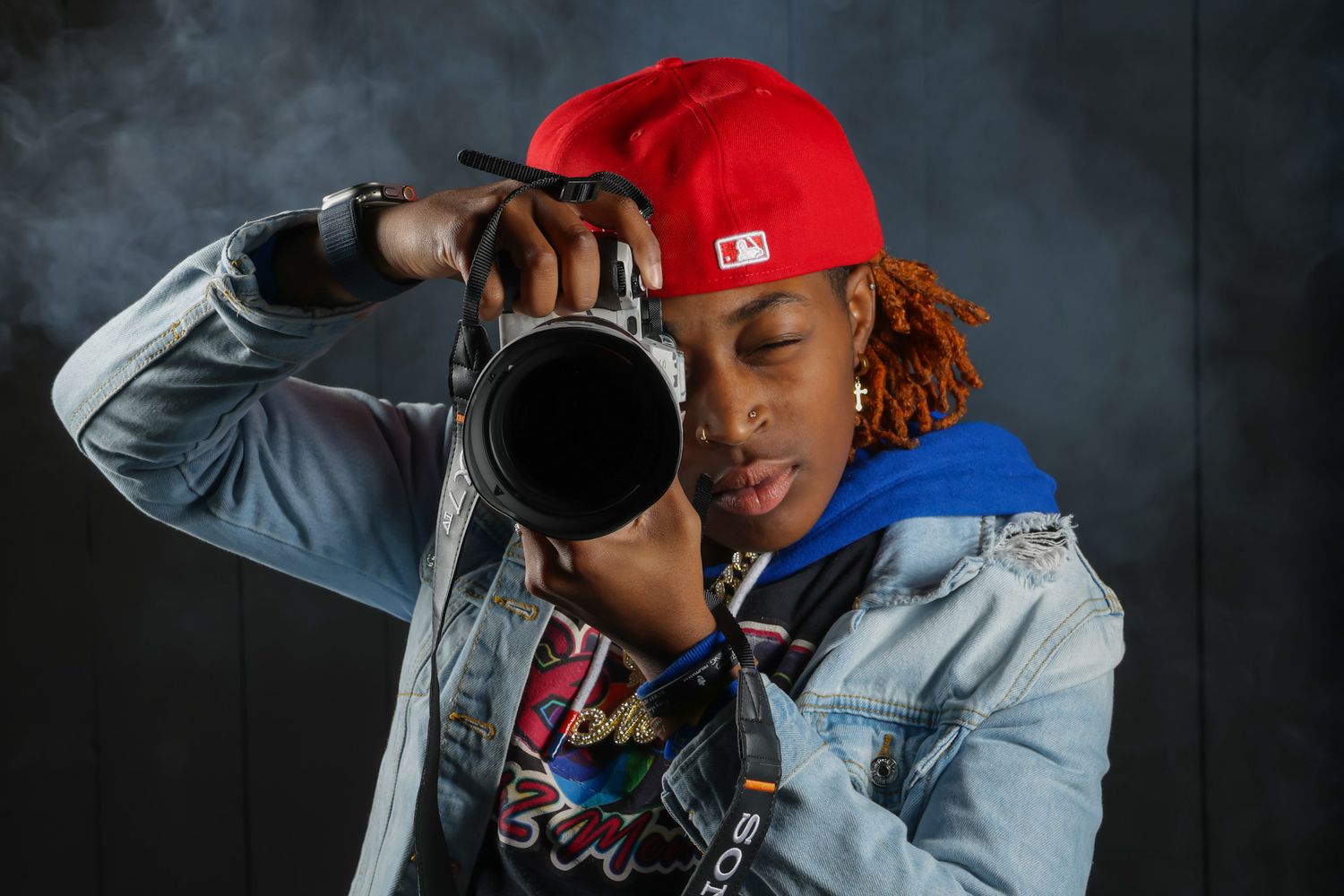 Young photographer in red cap aims DSLR camera on dark smoky background.