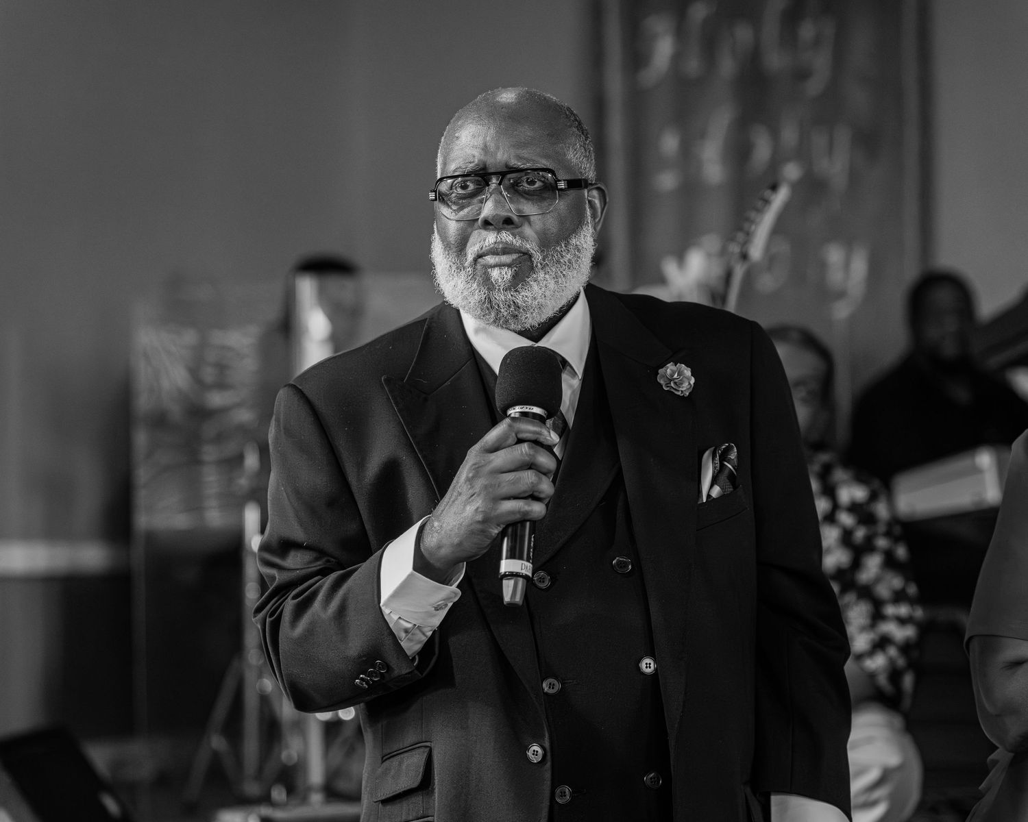Distinguished elderly man in suit speaking into microphone at formal black and white event.