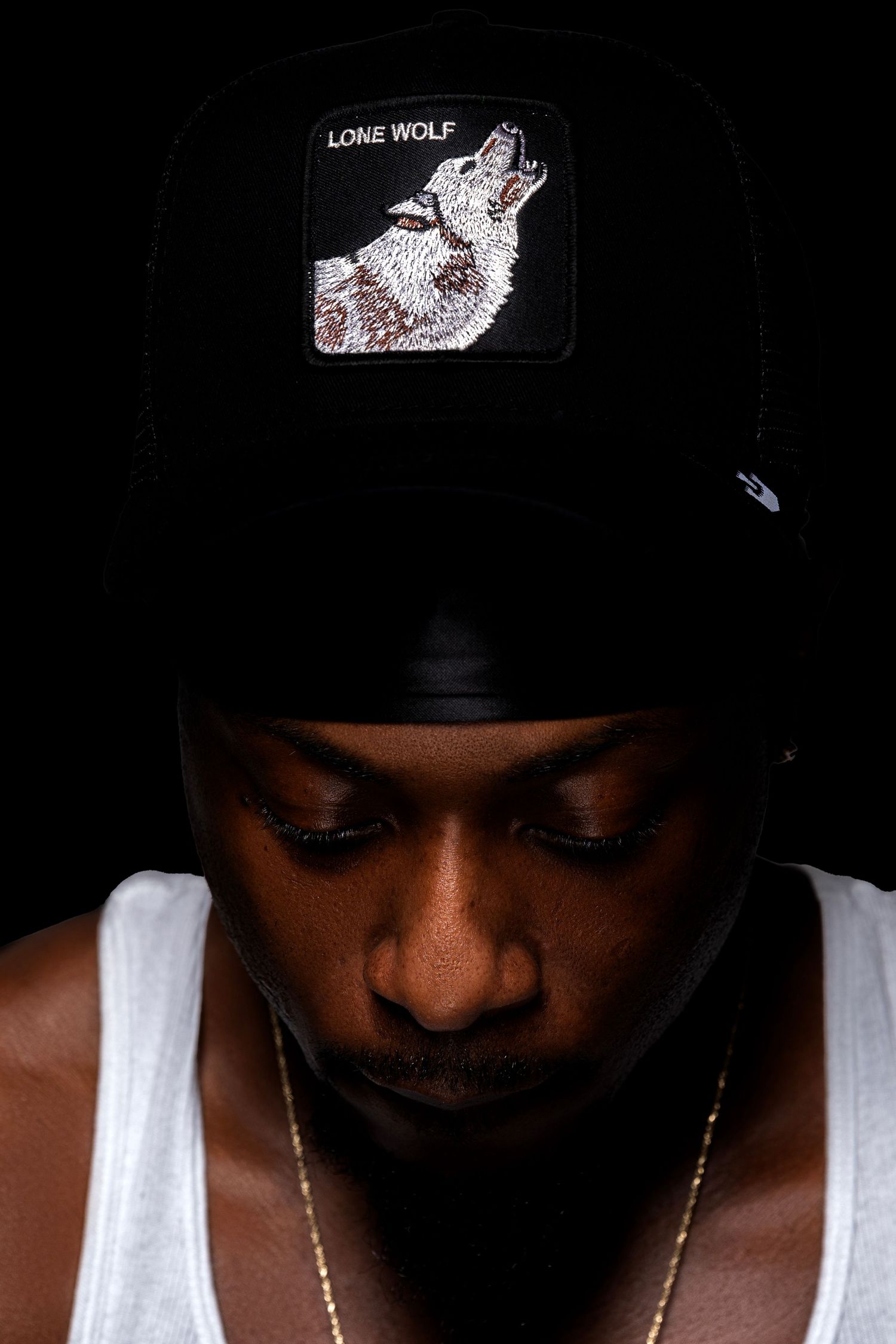 Man wearing a black Lone Wolf snapback cap looking down against dark background.